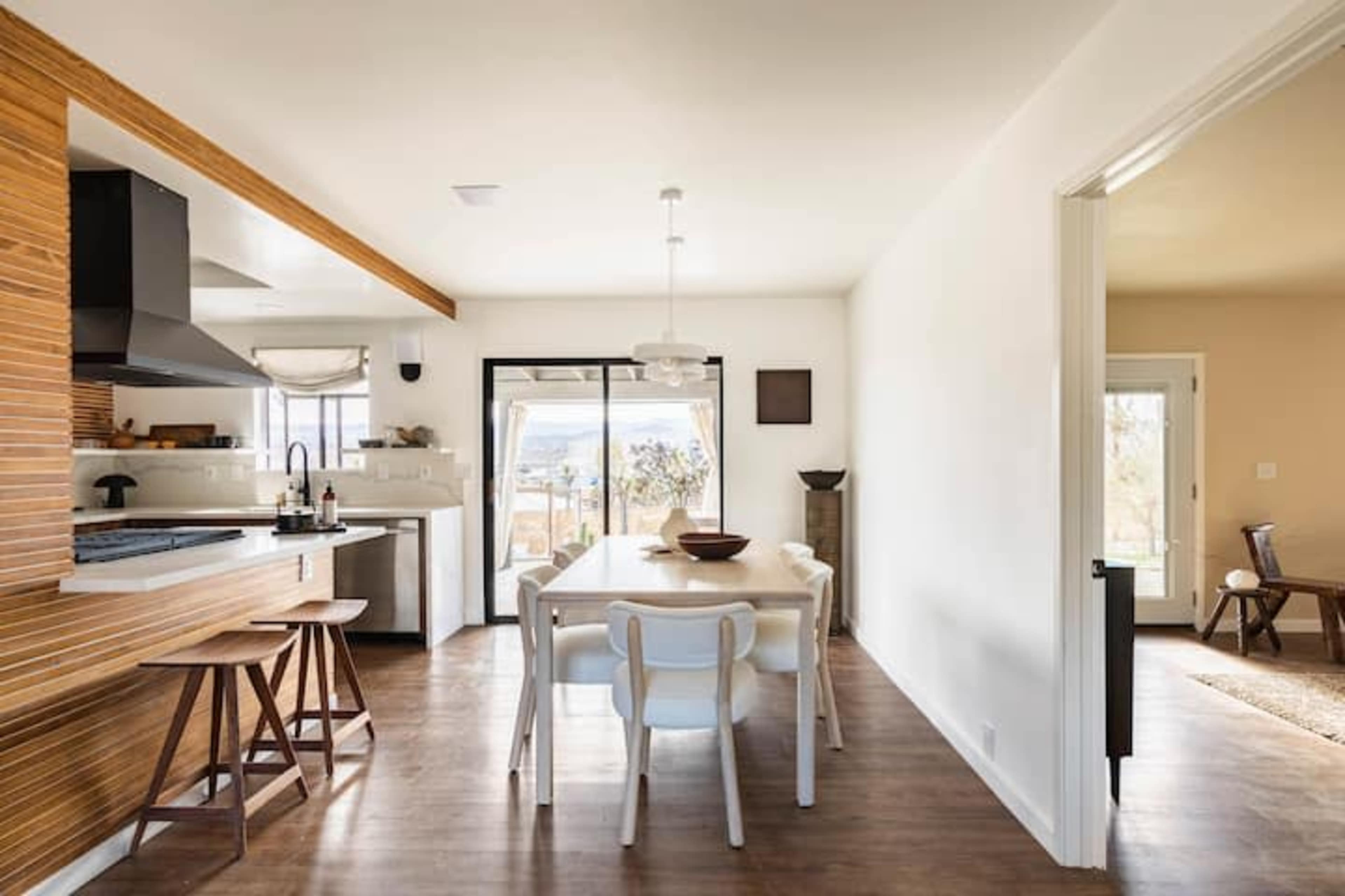 A modern kitchen and dining area feature a wooden table and white chairs, with large windows allowing natural light to enter.