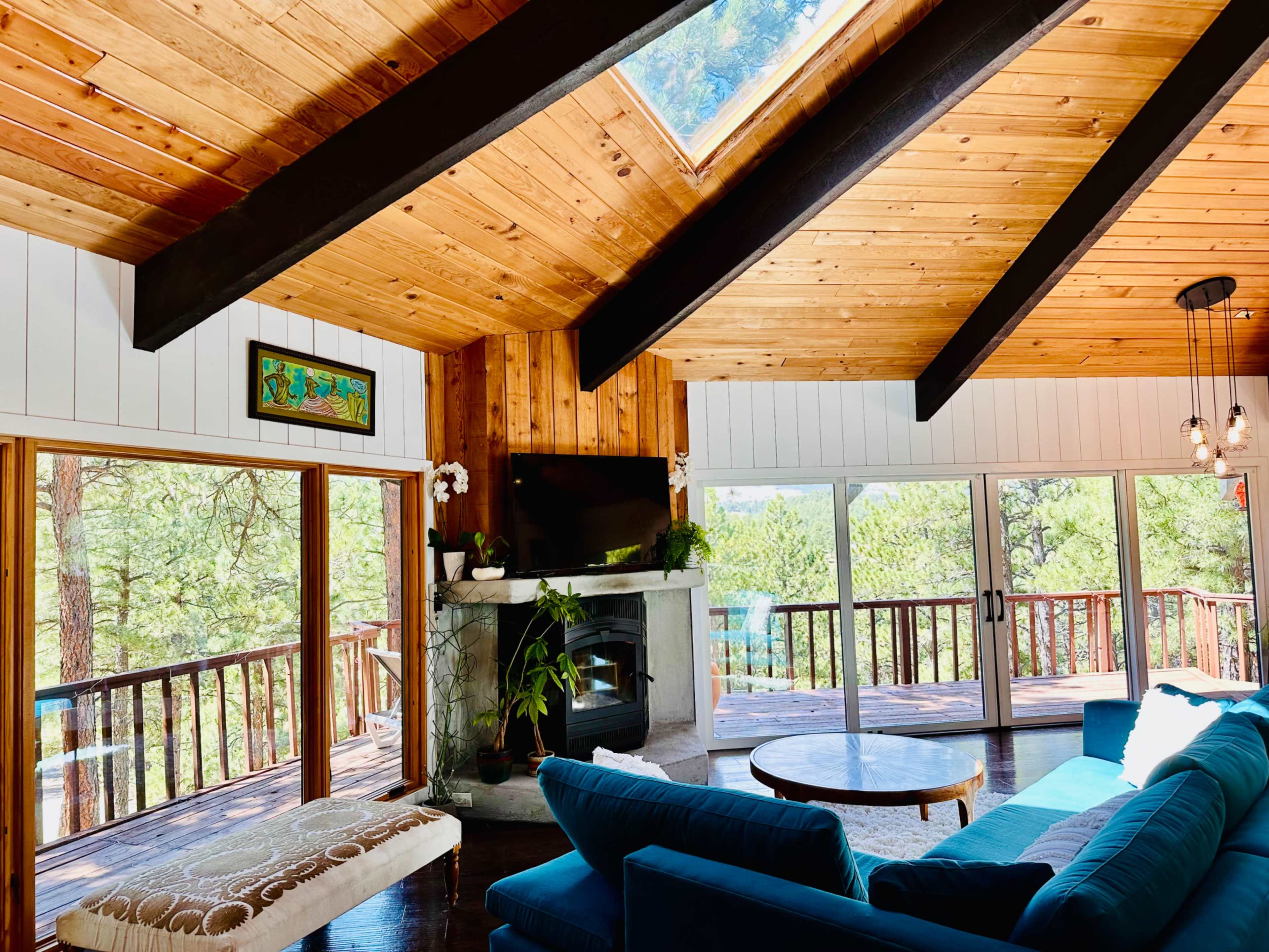 The image shows a bright, spacious living room with a wooden ceiling, large windows overlooking a forested area, and a modern fireplace.