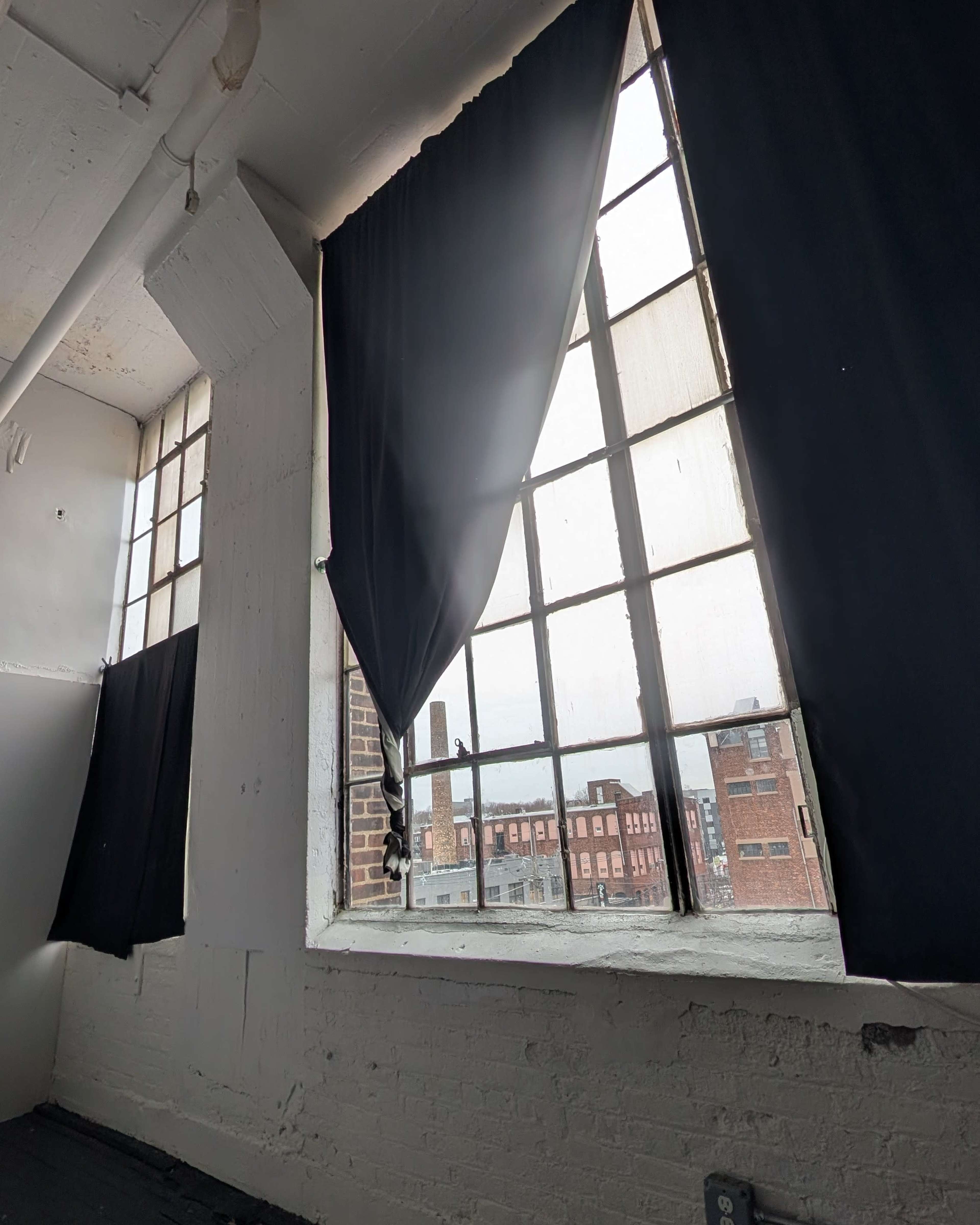 The image shows a room with large windows covered by black curtains, revealing a view of neighboring buildings in an industrial setting.