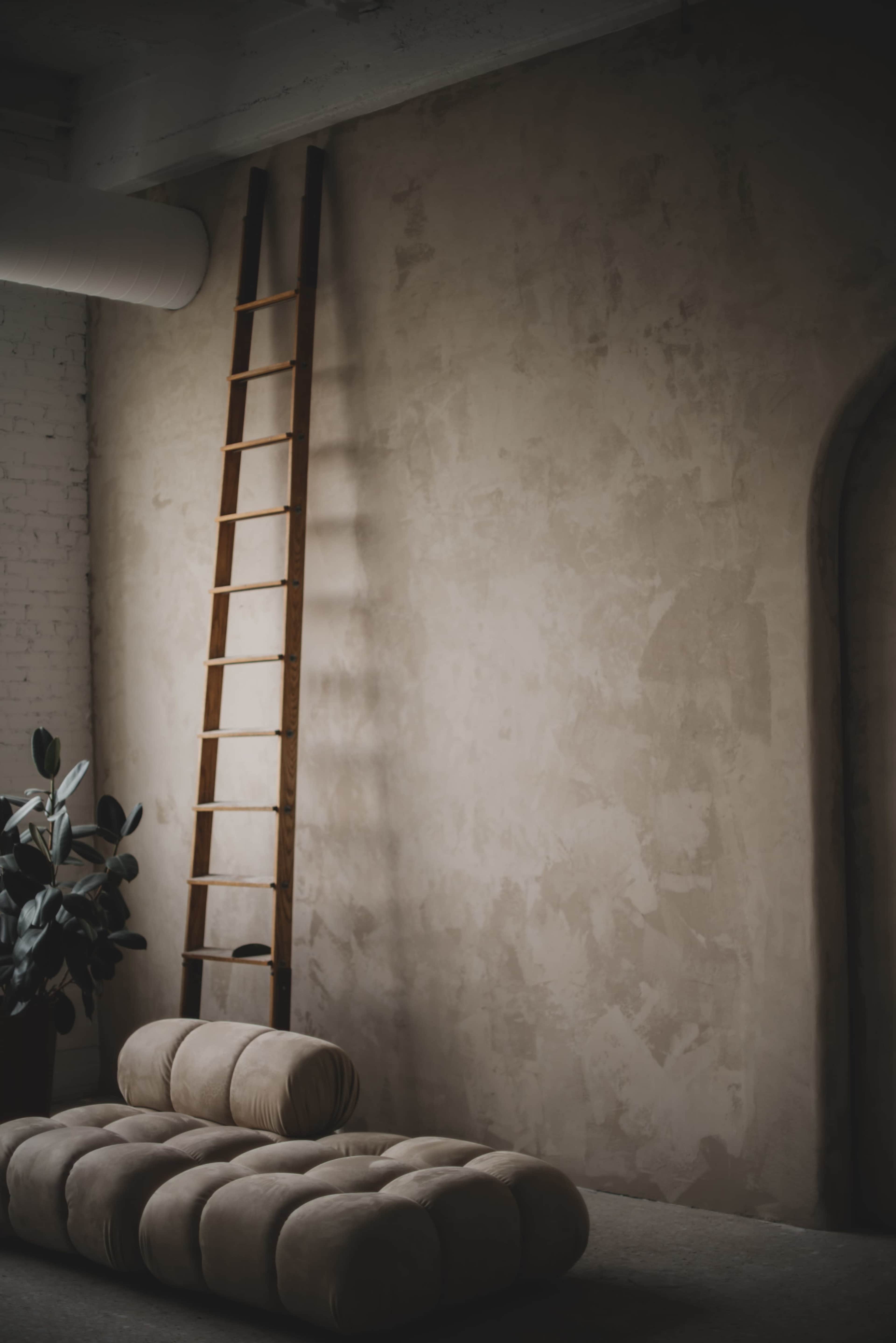 A wooden ladder leans against a textured wall beside a low, padded sofa in a dimly lit room.