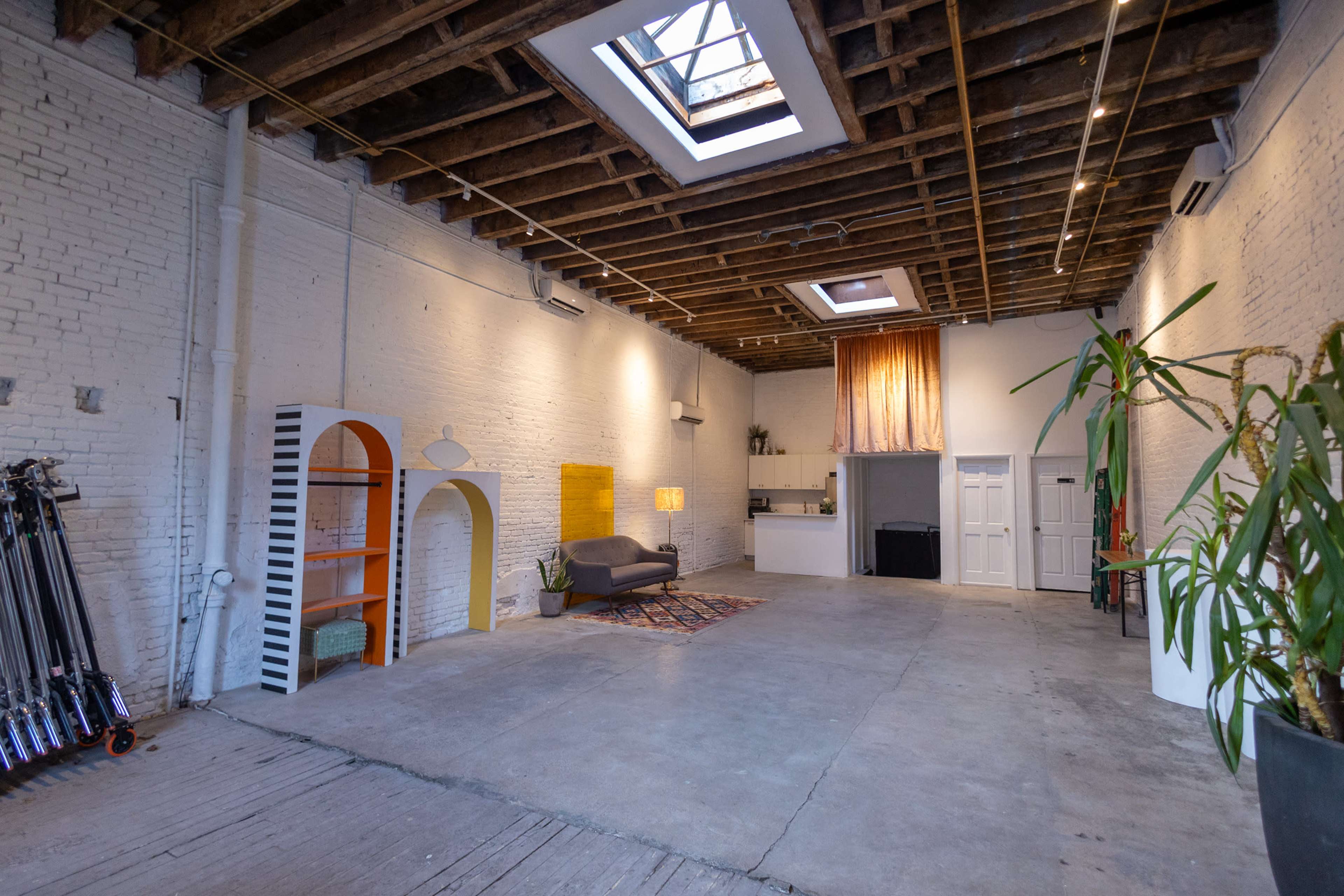 A spacious, industrial-style room with exposed wooden beams, a gray sofa, and a kitchenette, surrounded by white brick walls.