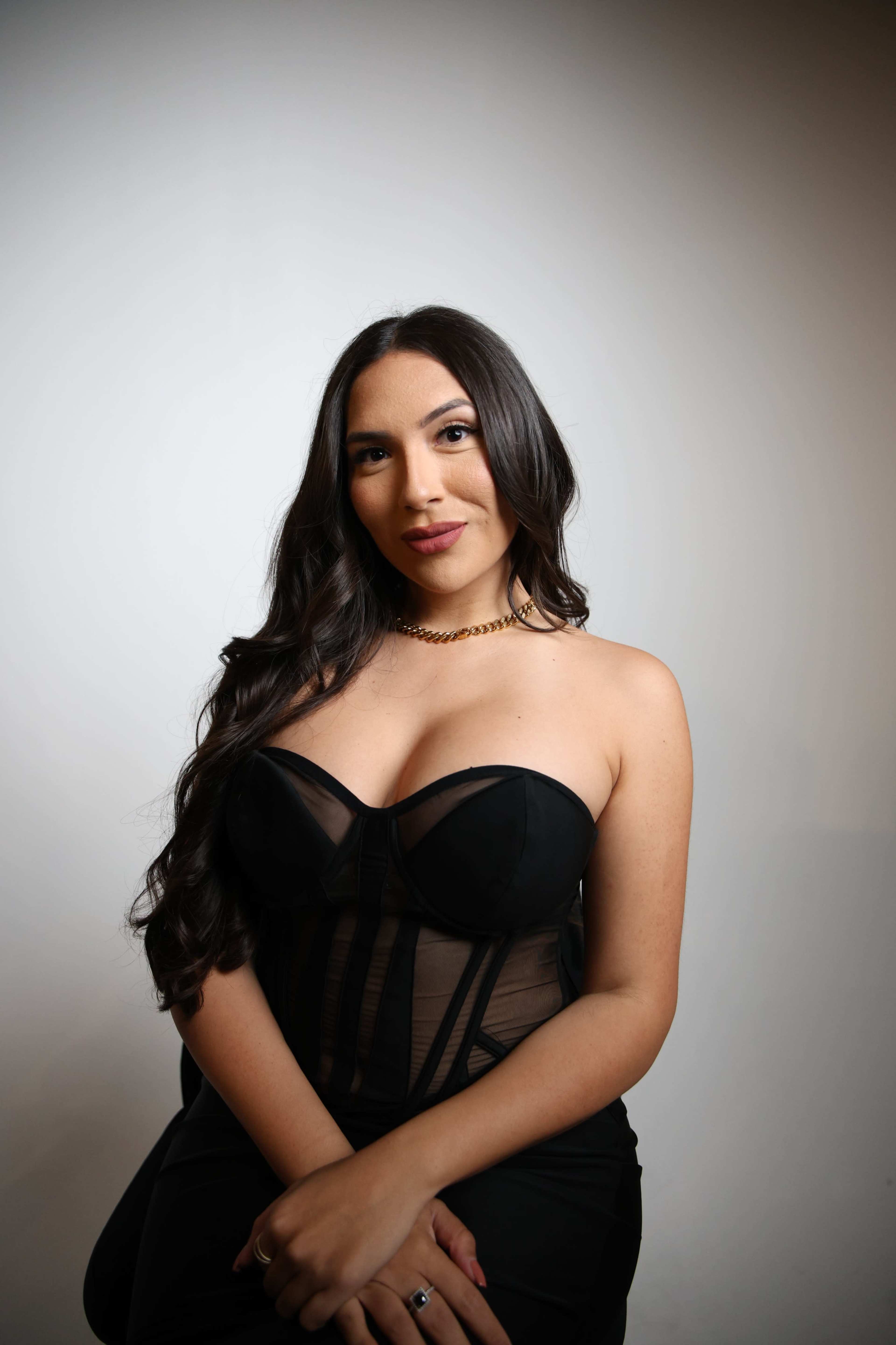 A woman with long dark hair poses sitting down, wearing a black strapless top and a gold chain necklace against a plain background.
