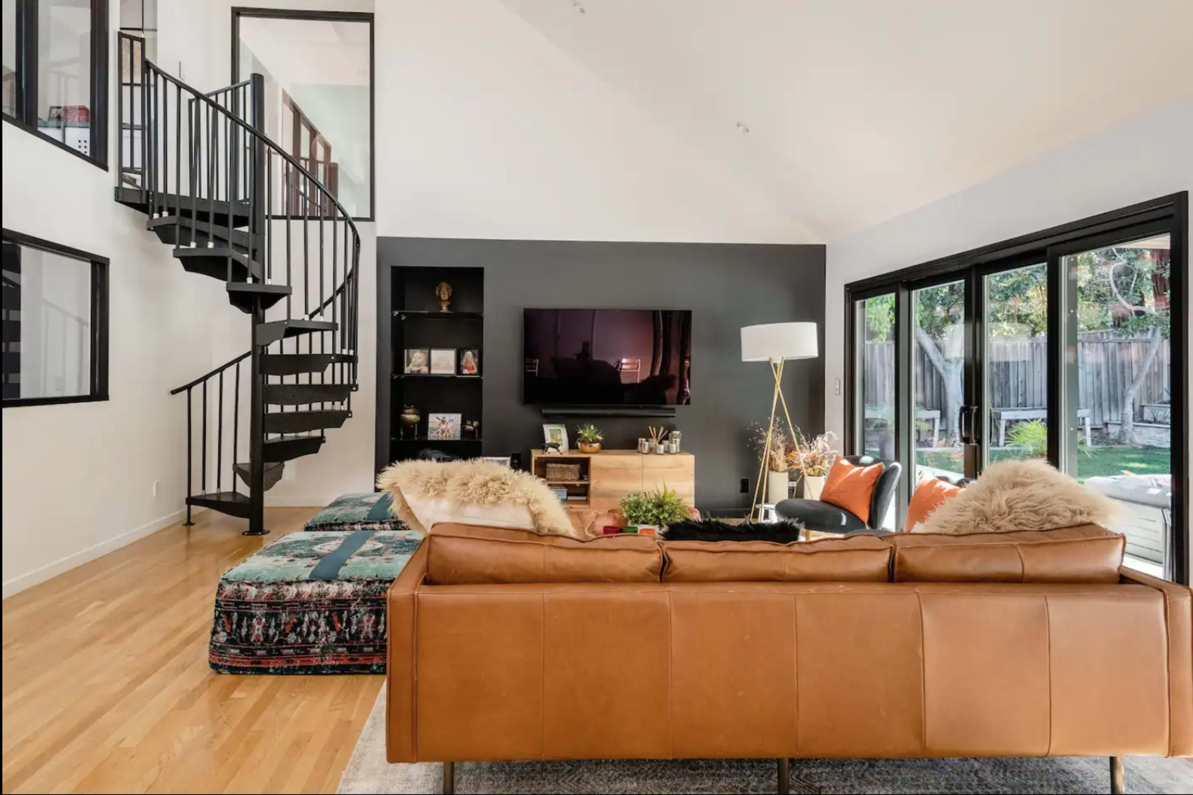 A modern living room features a leather sofa, a flat-screen TV on a dark accent wall, and large windows overlooking a garden, with a spiral staircase in the background.
