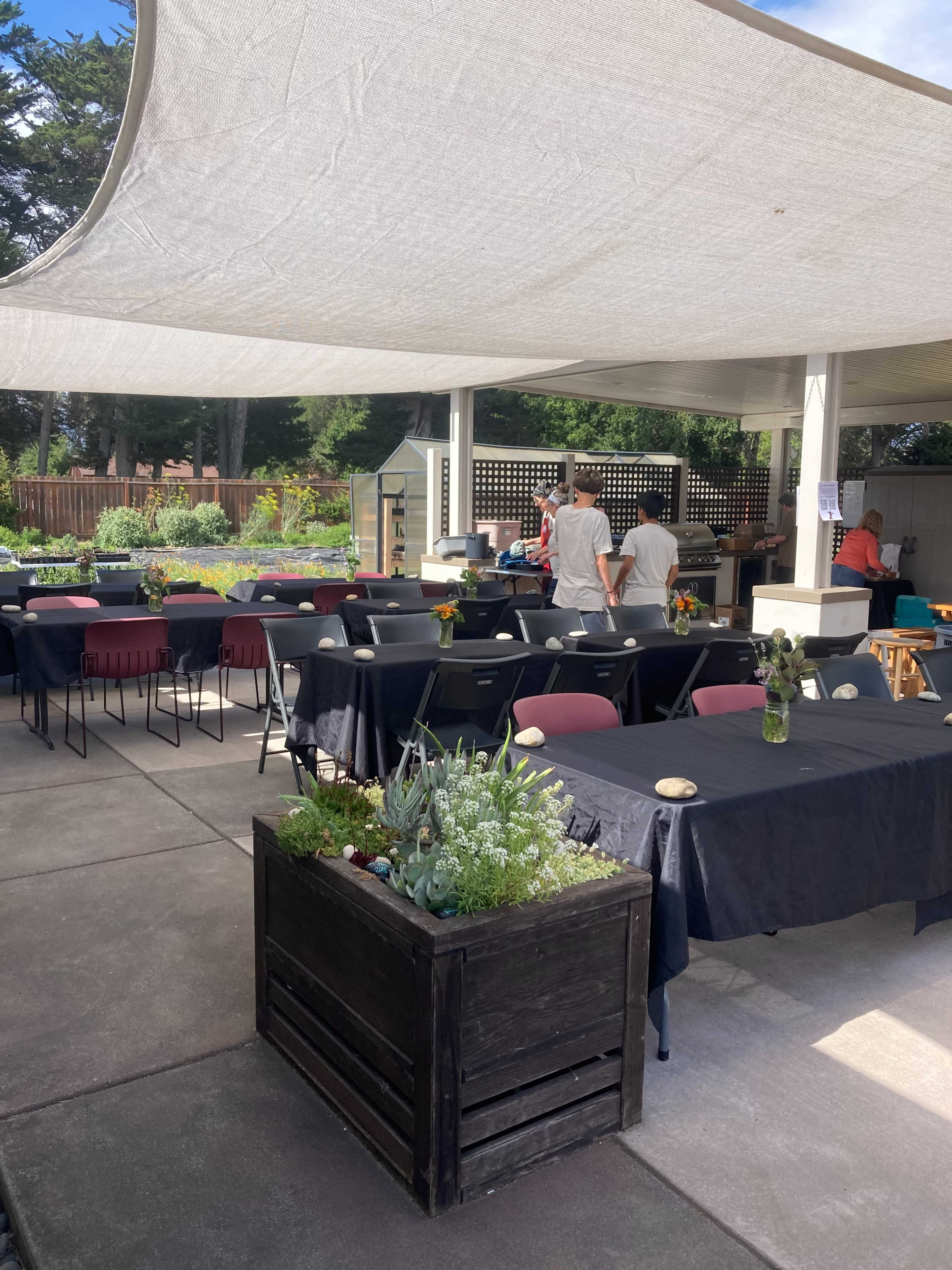 A shaded outdoor dining area is set up with several tables and chairs, while people prepare food in the background.