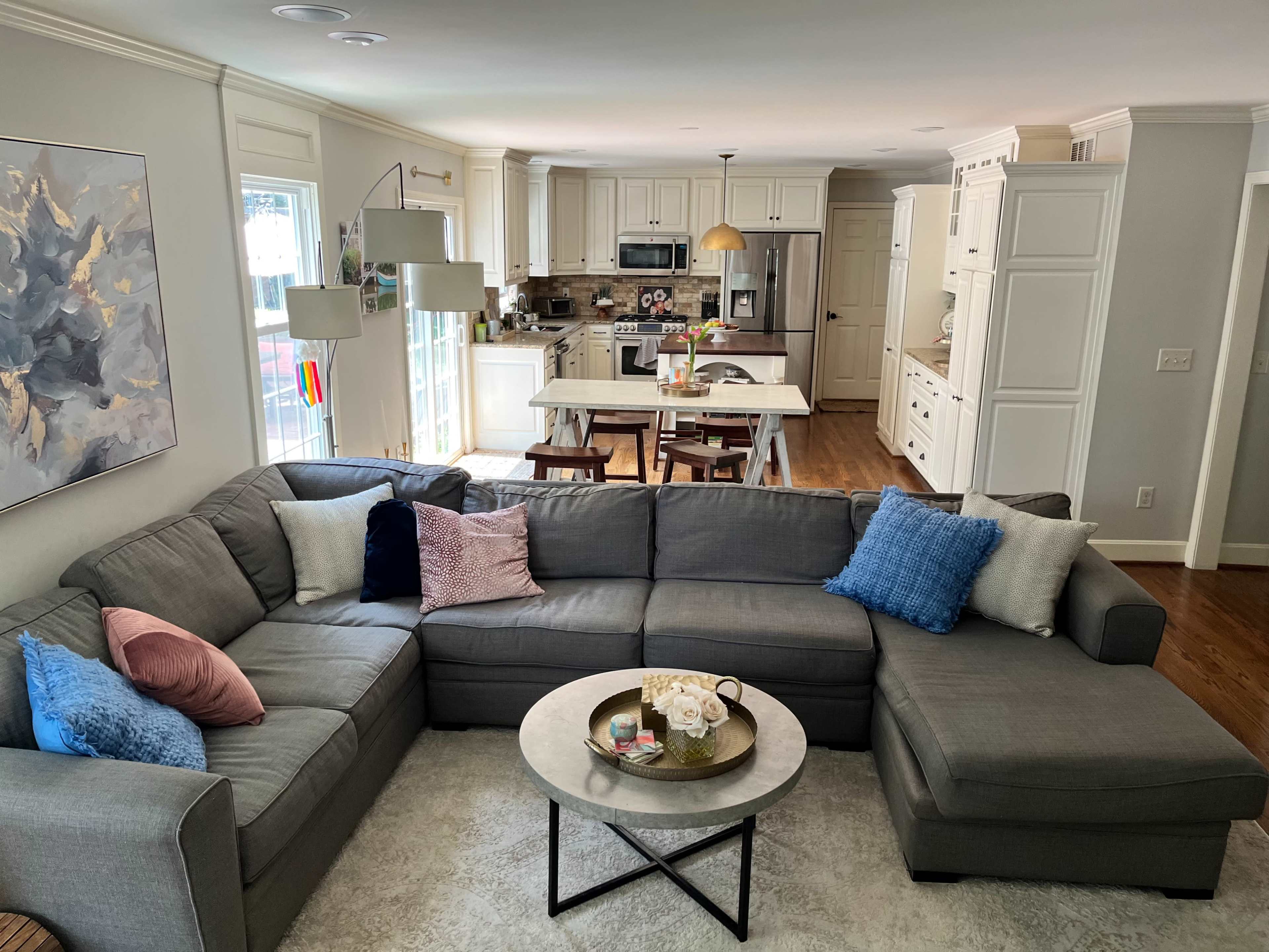 A spacious living room features a large gray sectional sofa with colorful throw pillows and a circular coffee table, overlooking an open kitchen with white cabinetry.