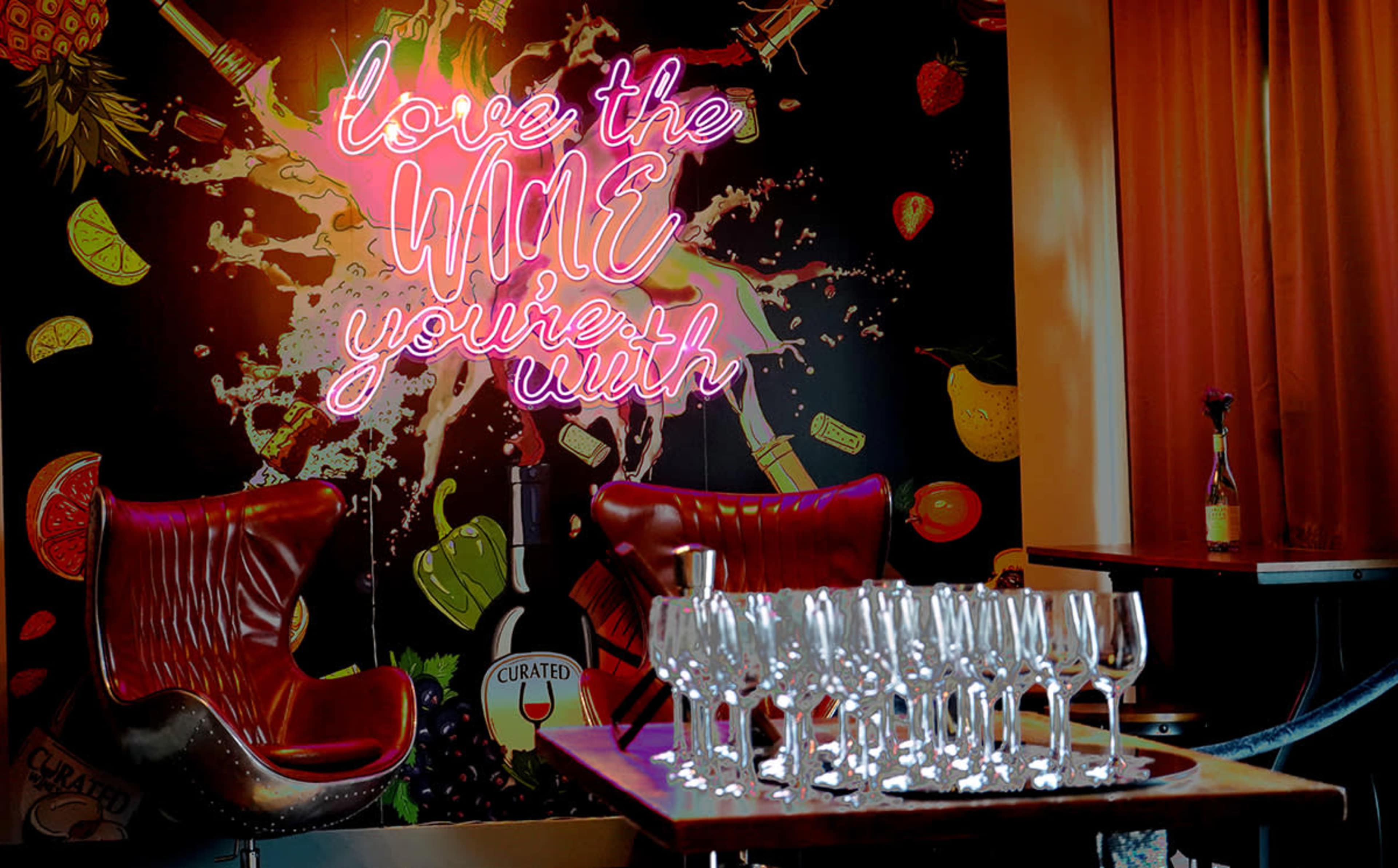 The image shows a vibrant bar scene with a neon sign that reads "love the wine you're with" against a colorful backdrop, featuring two distinctive red chairs and a row of sparkling wine glasses on a table.