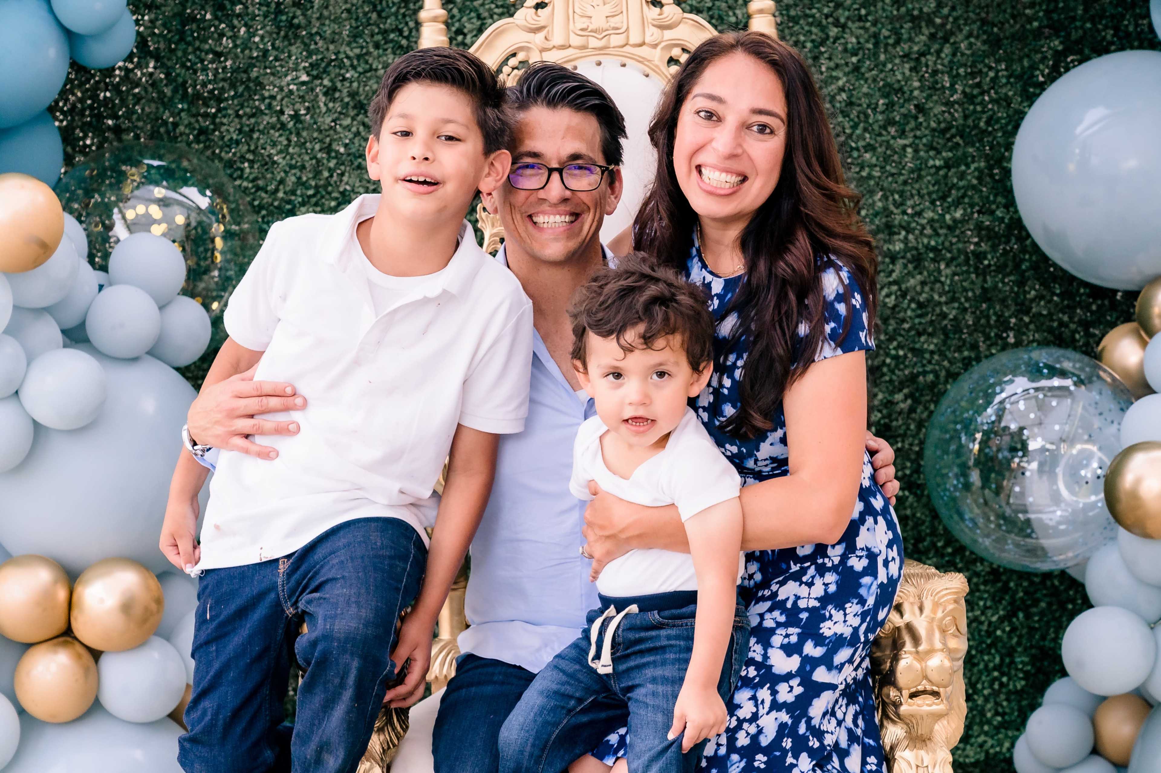 A family of four is smiling together while seated in front of a backdrop of greenery and decorative balloons.