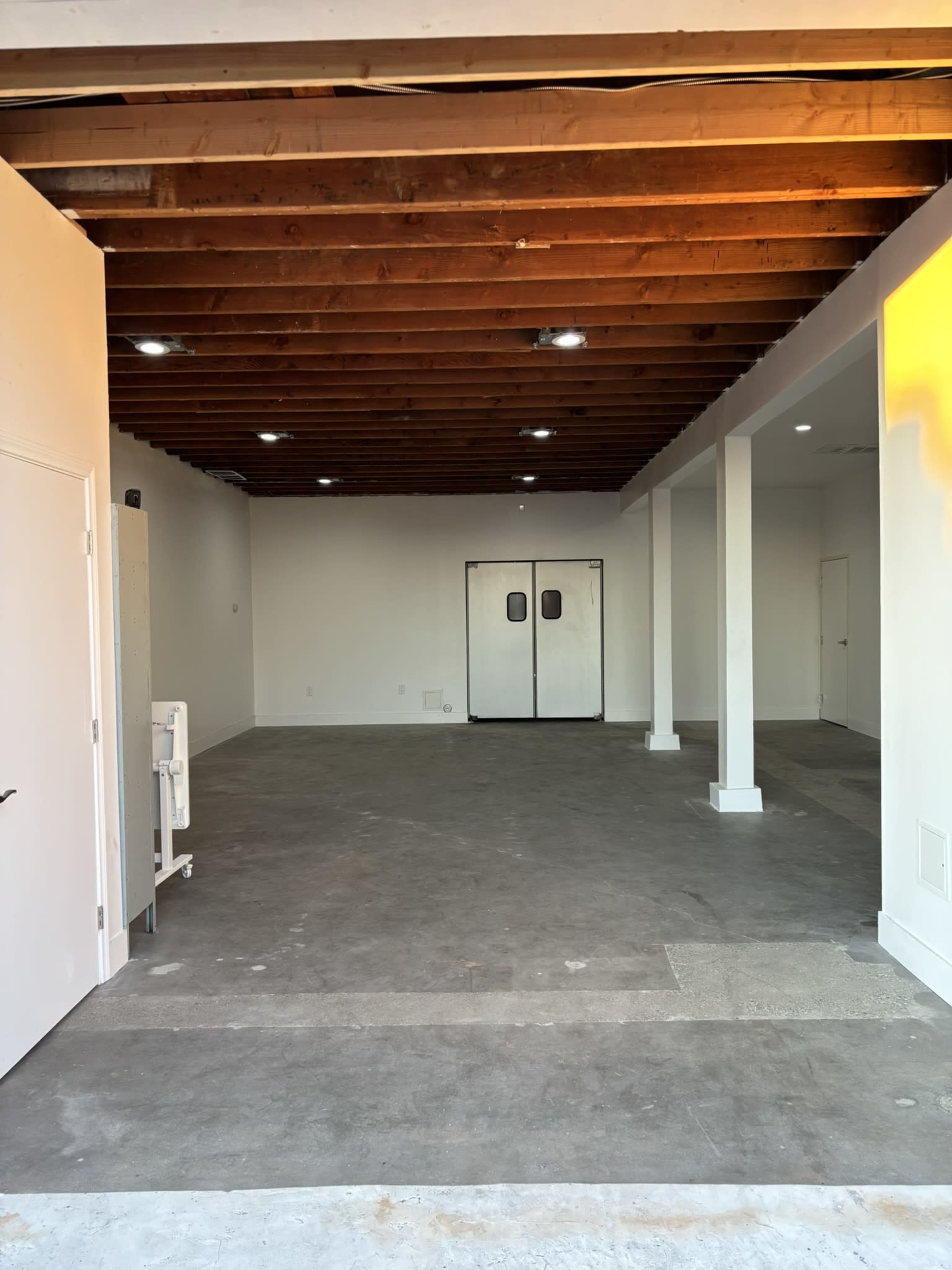 The image shows an empty interior space with exposed wooden beams and concrete flooring, featuring white walls and two double doors at the far end.