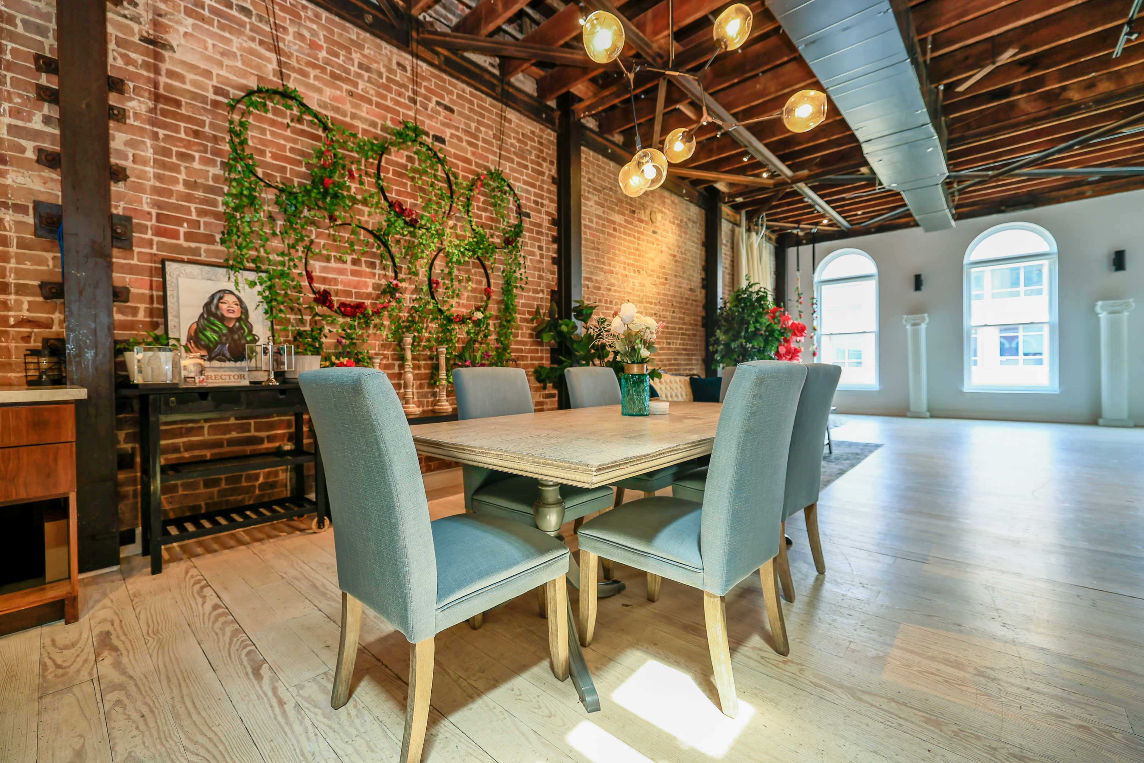 A dining area features a wooden table surrounded by upholstered chairs, with a brick wall adorned with greenery and pendant lights above.