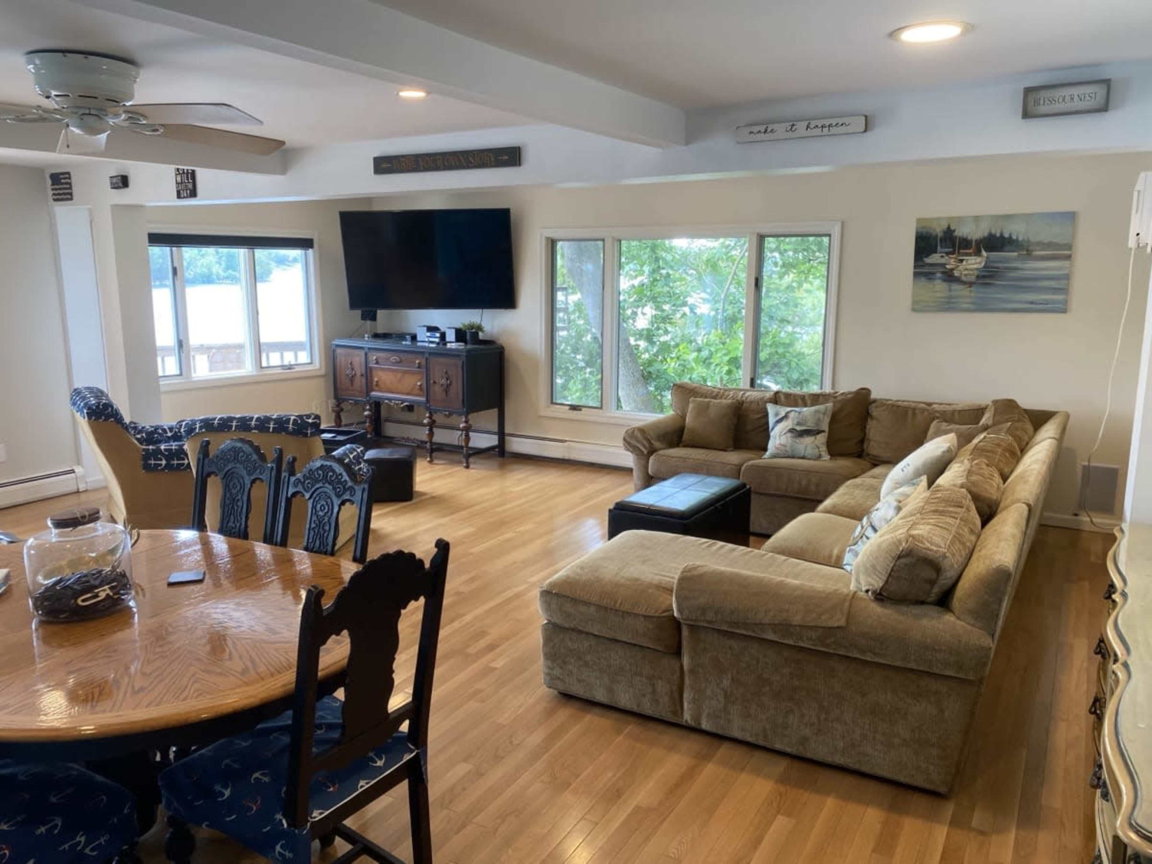 A spacious living room with a large sectional sofa, a coffee table, a dining table, and several windows overlooking a natural landscape.