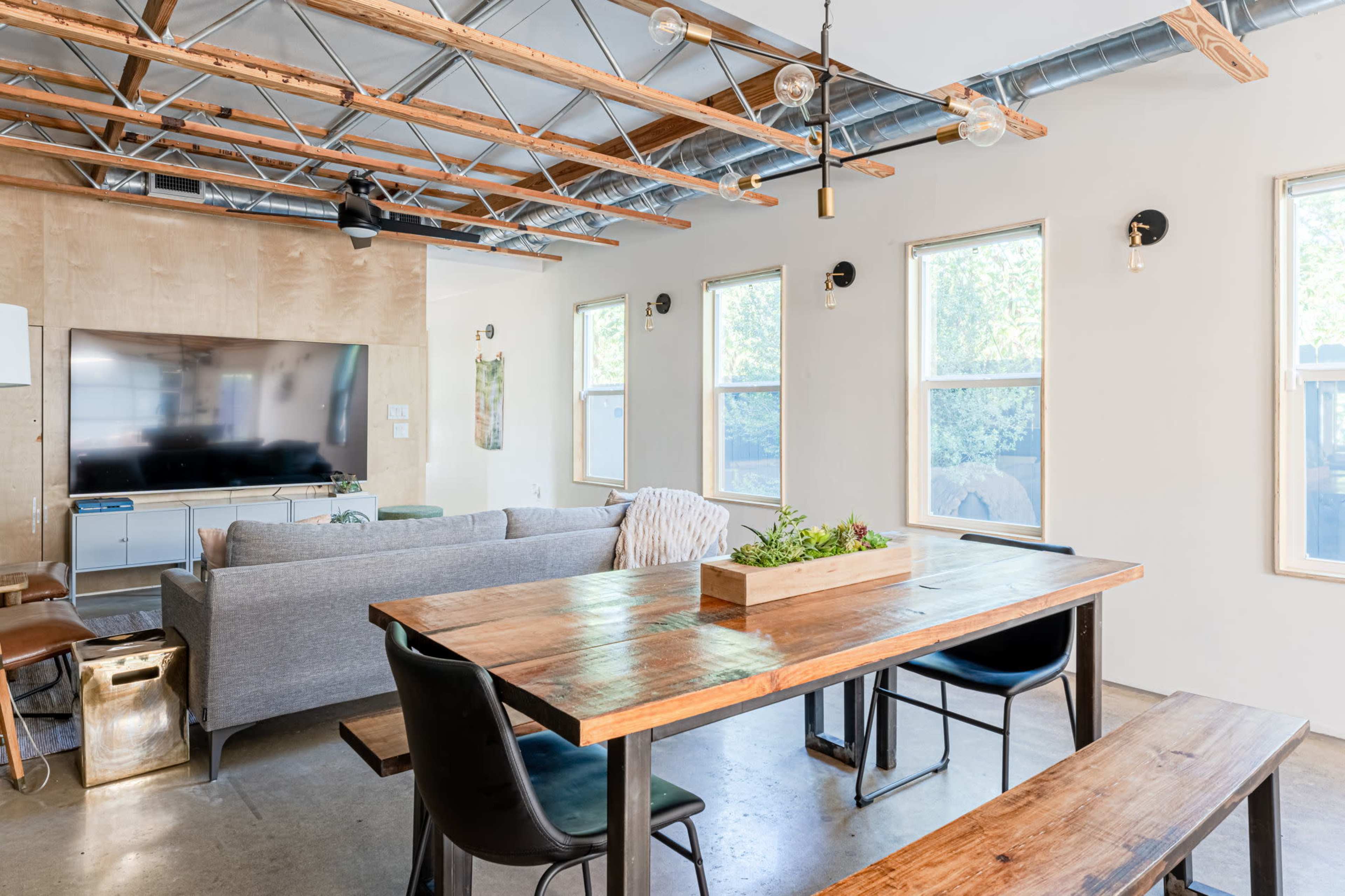 The image shows a modern living and dining area with a wooden dining table, chairs, and a comfortable sofa, set beneath a ceiling with exposed beams and hanging lights.