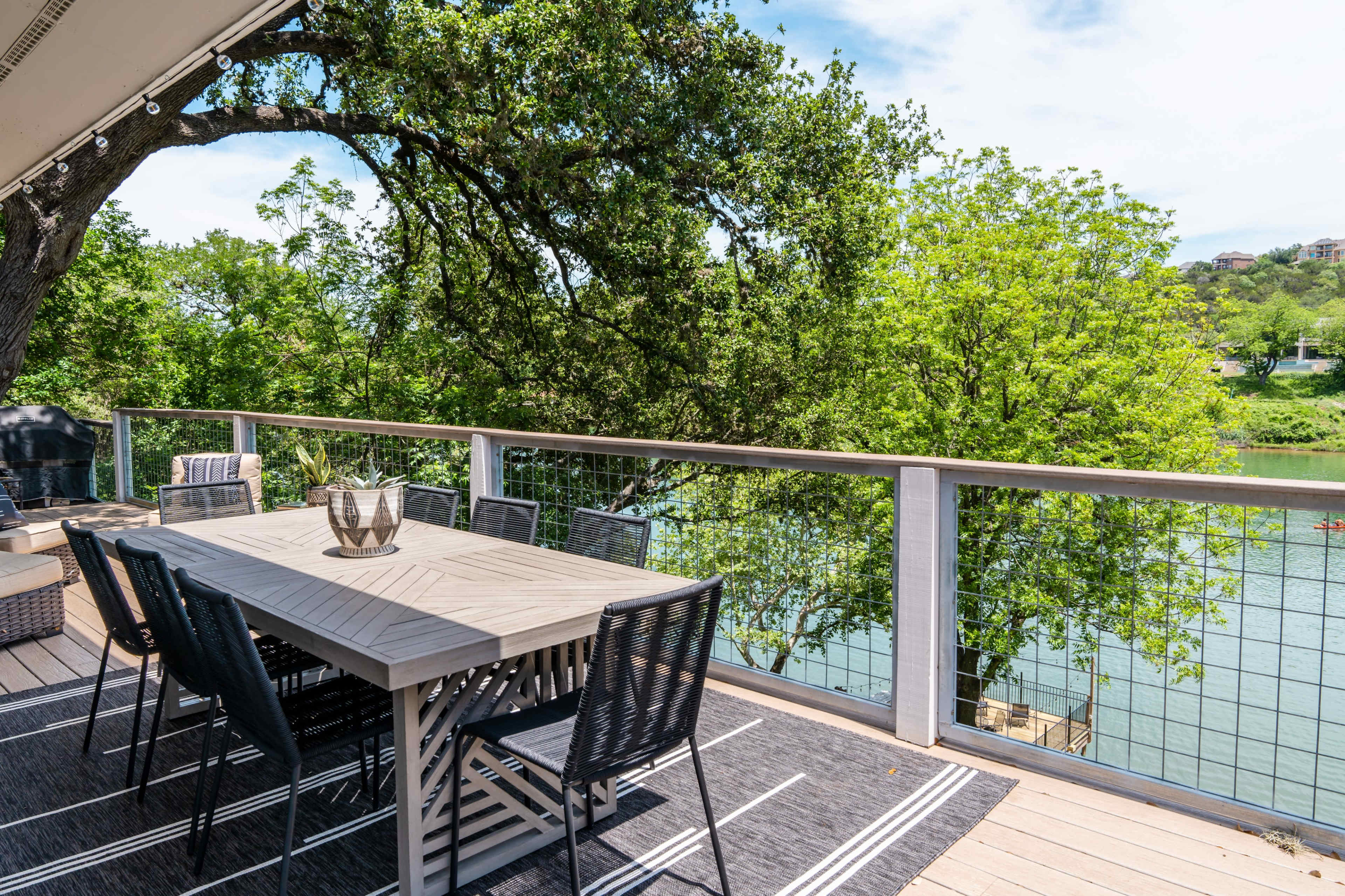 A modern outdoor dining area features a rectangular table surrounded by chairs on a deck overlooking a river and lush greenery.