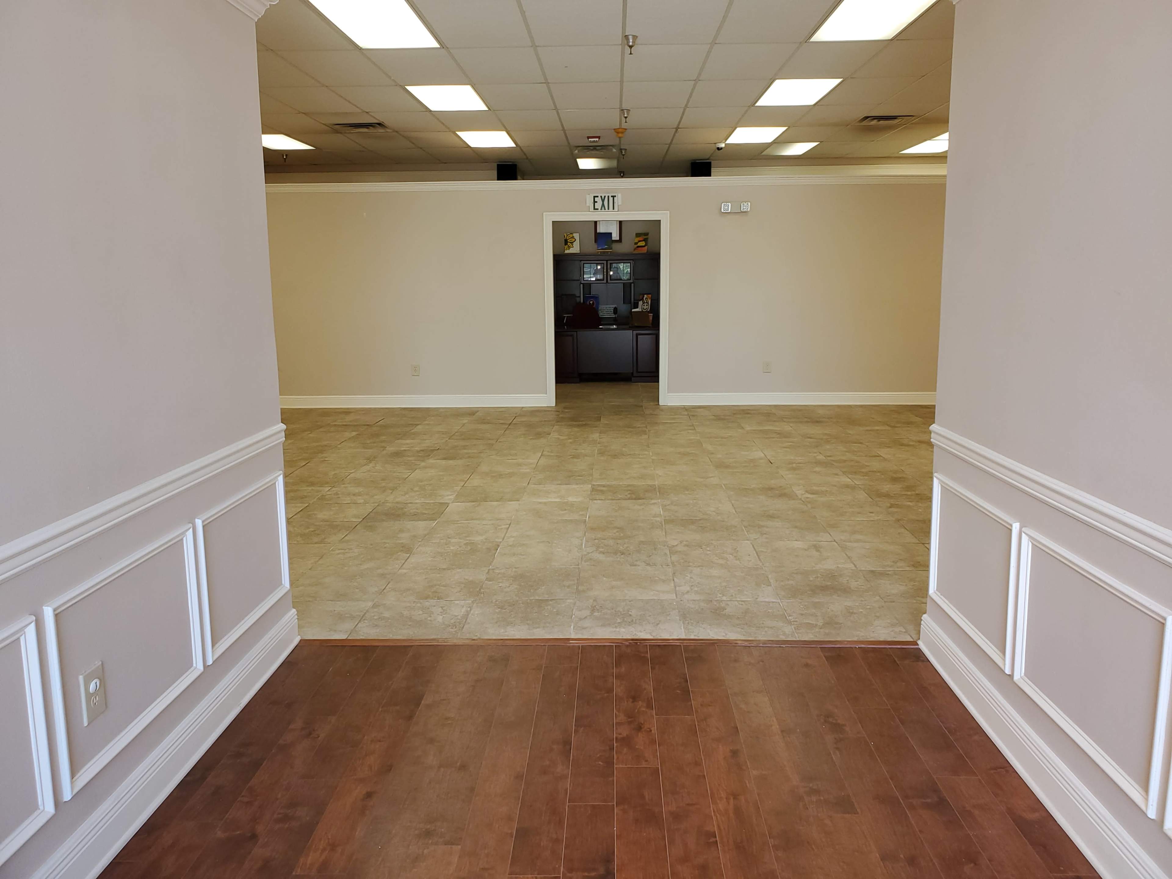 The image shows a spacious, empty room with tiled flooring and a doorway leading to another area in the background.