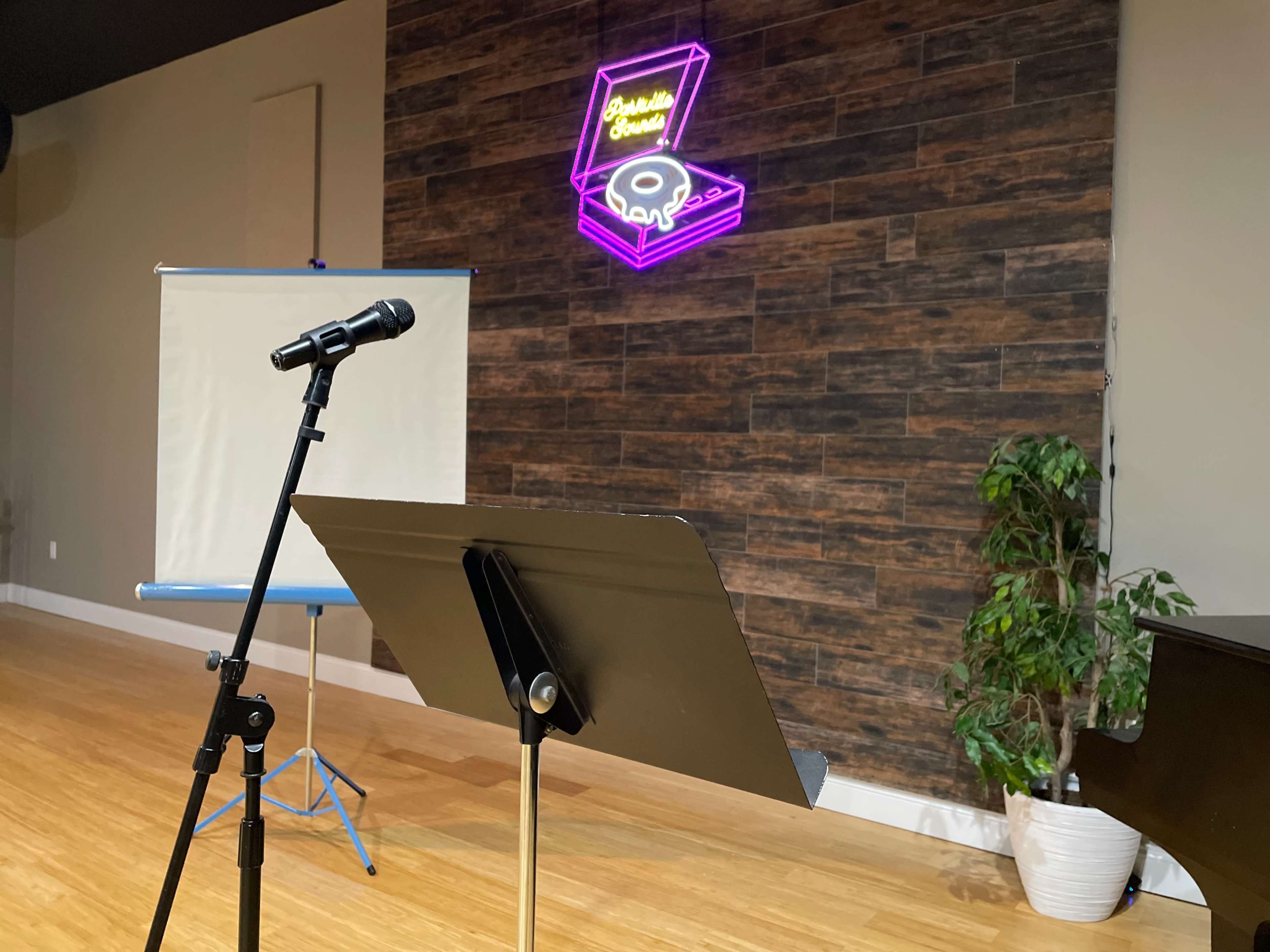 A microphone stands ready on a stand in front of a projector screen, with a neon sign featuring a record player on the wall behind it.