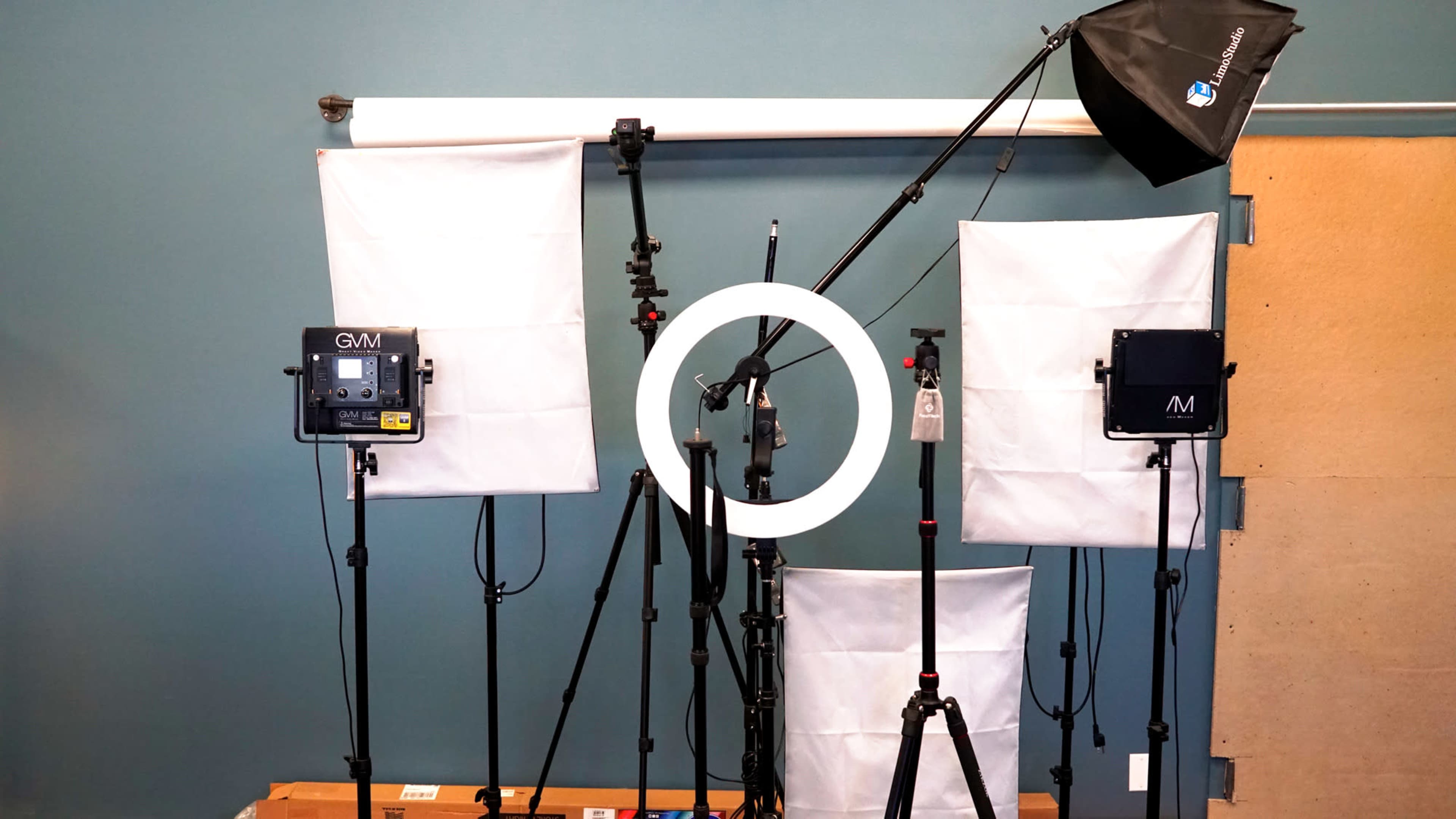 The image shows a setup of various photography lights, including a ring light and two softbox lights, positioned on tripods against a teal-painted wall.