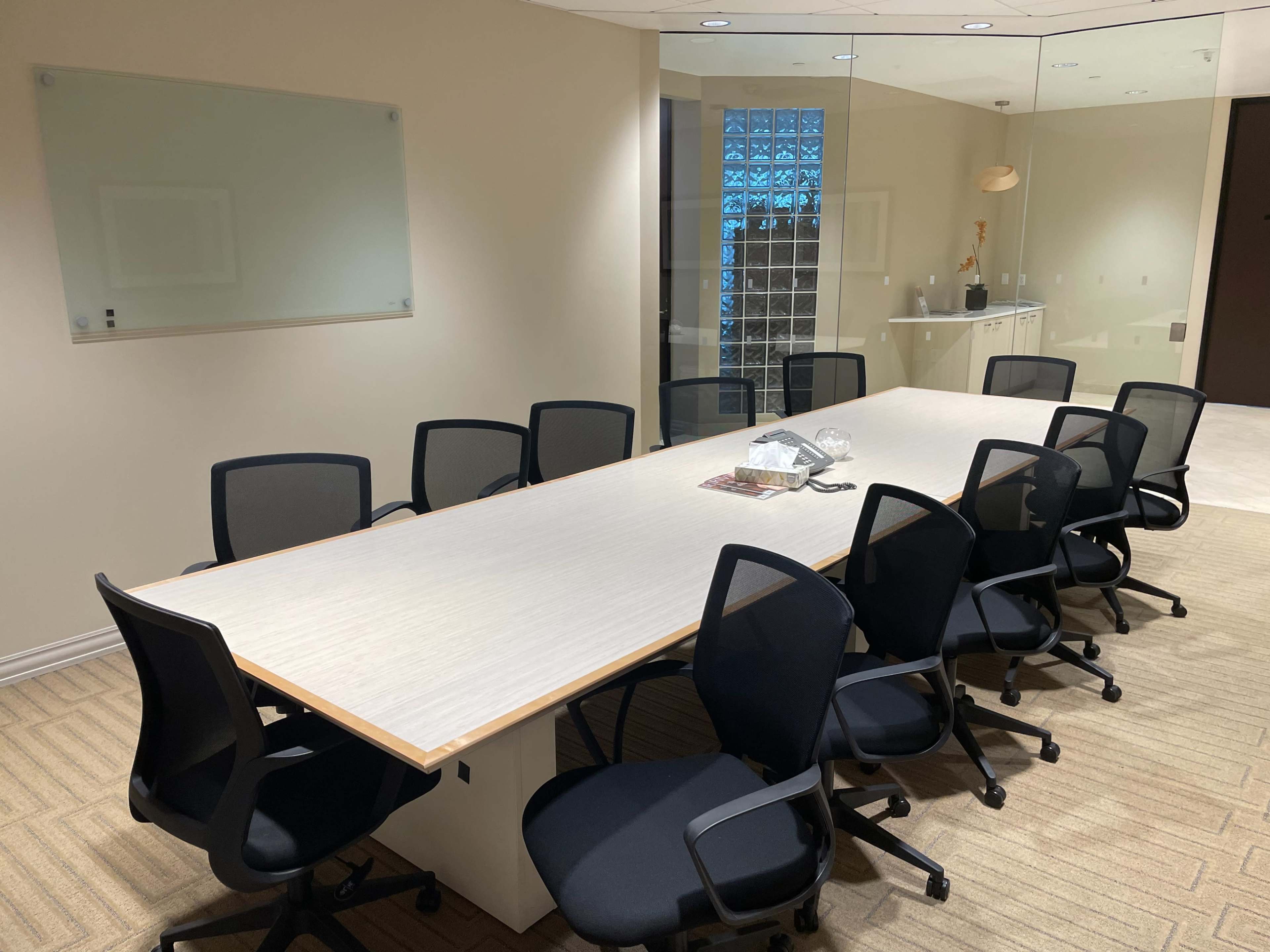 A large conference room features a long table surrounded by black chairs and a wall with glass blocks.