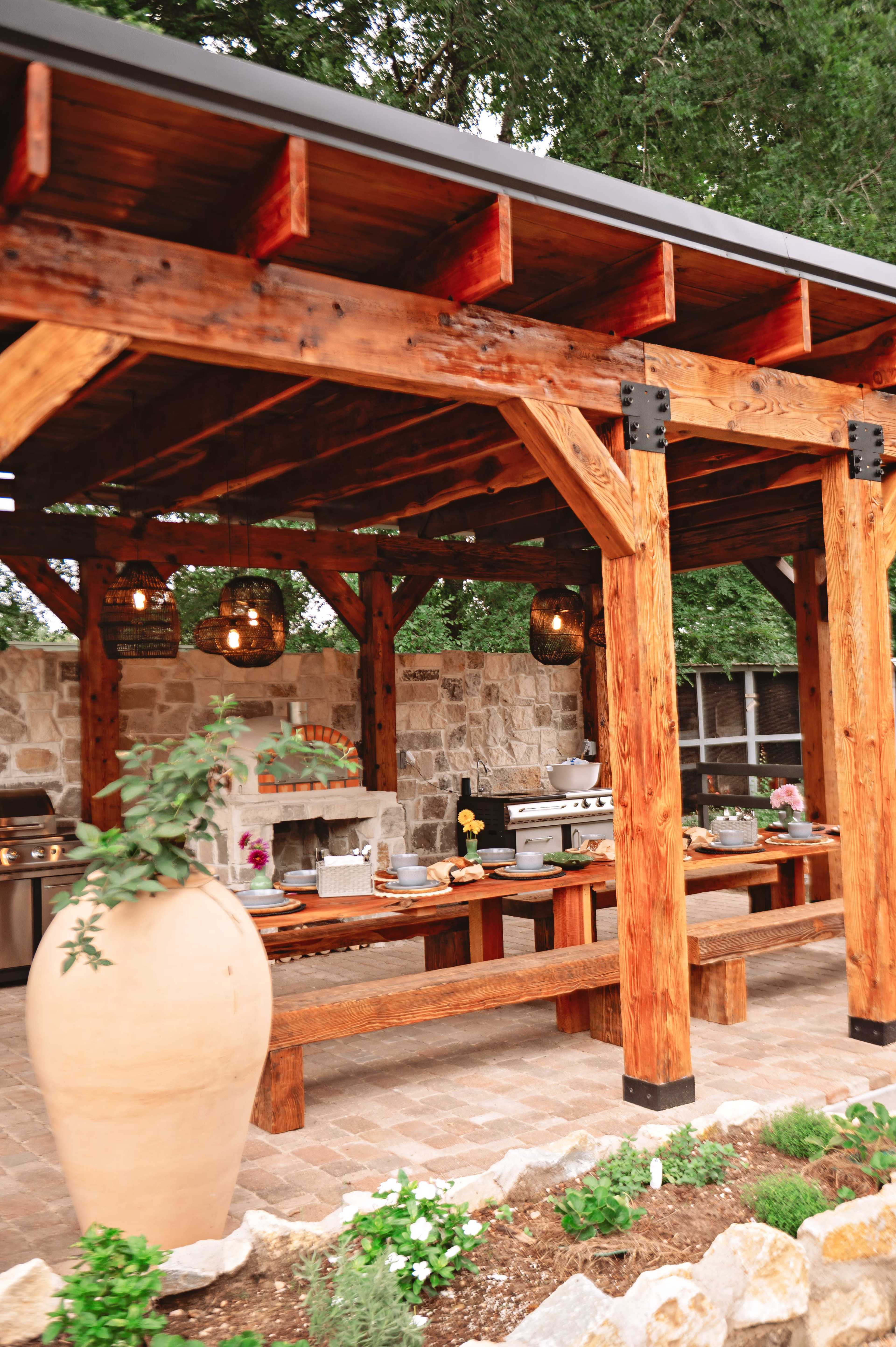 The image shows an outdoor kitchen area with a wooden pergola, a stone wall, a large clay planter, and a long wooden dining table.