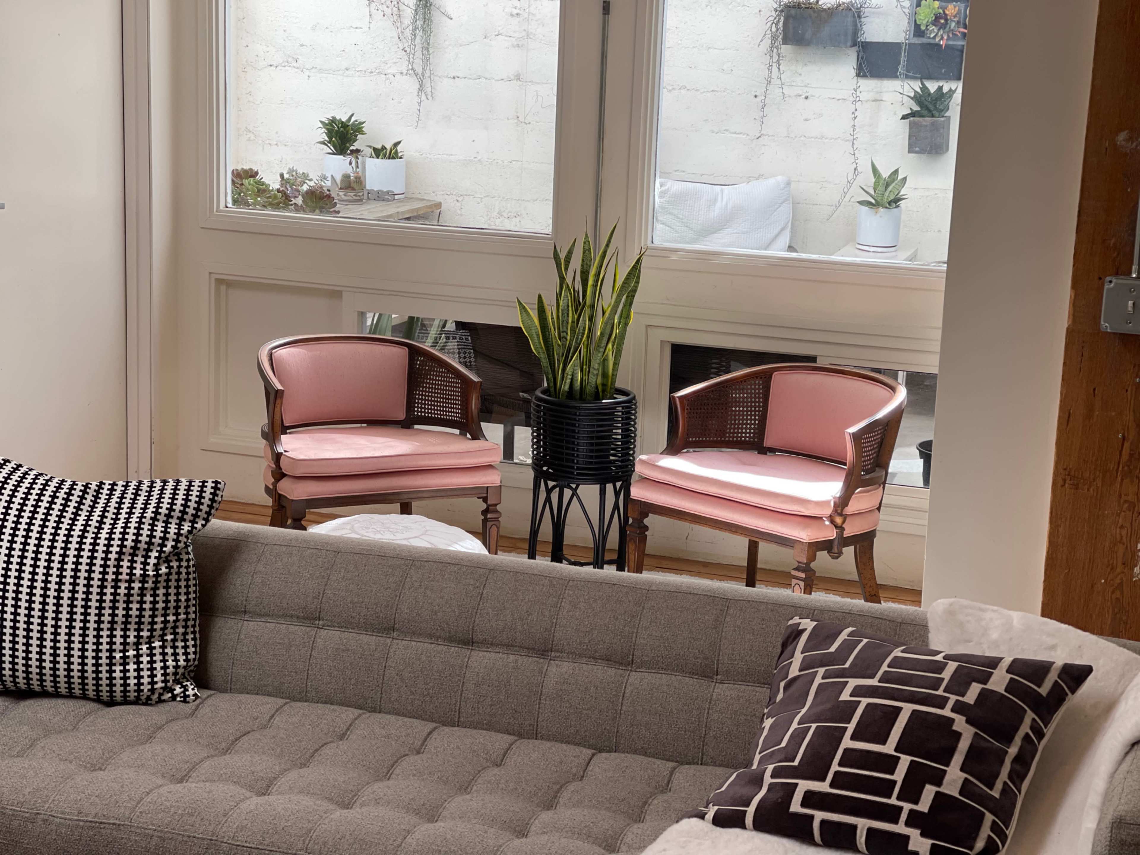 A cozy seating area featuring a gray couch, two pink upholstered chairs, and a potted plant positioned near a large window.