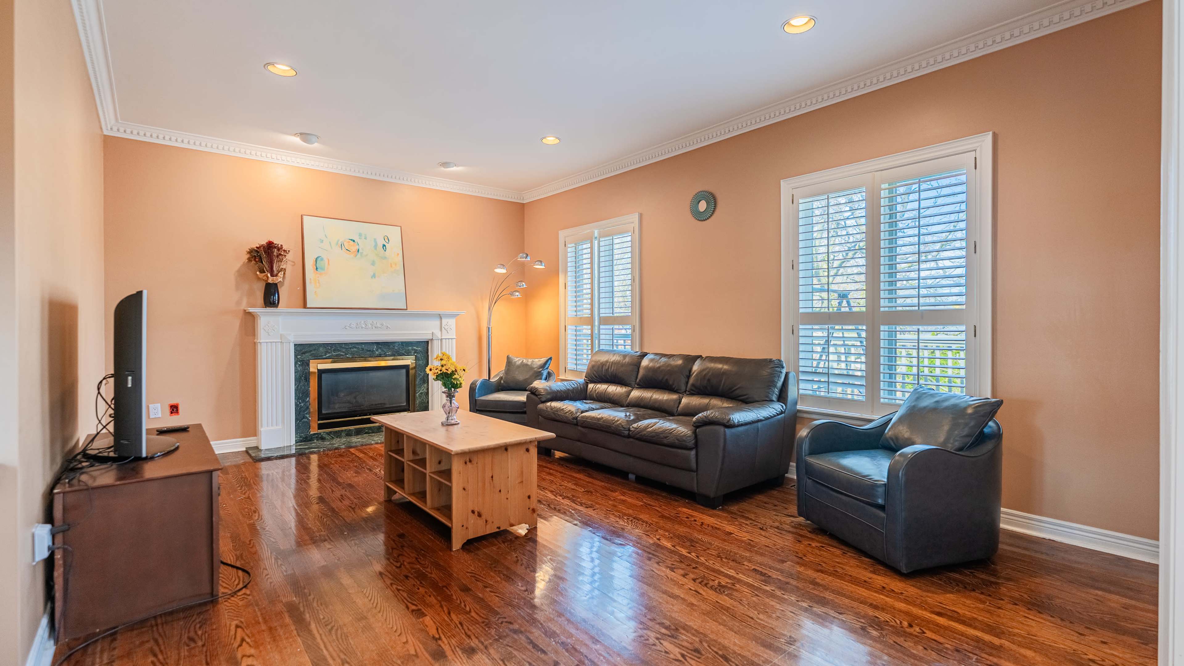 The image shows a living room with a leather sofa, an armchair, a wooden coffee table, and a fireplace, all set against light-colored walls and bright natural light from the windows.