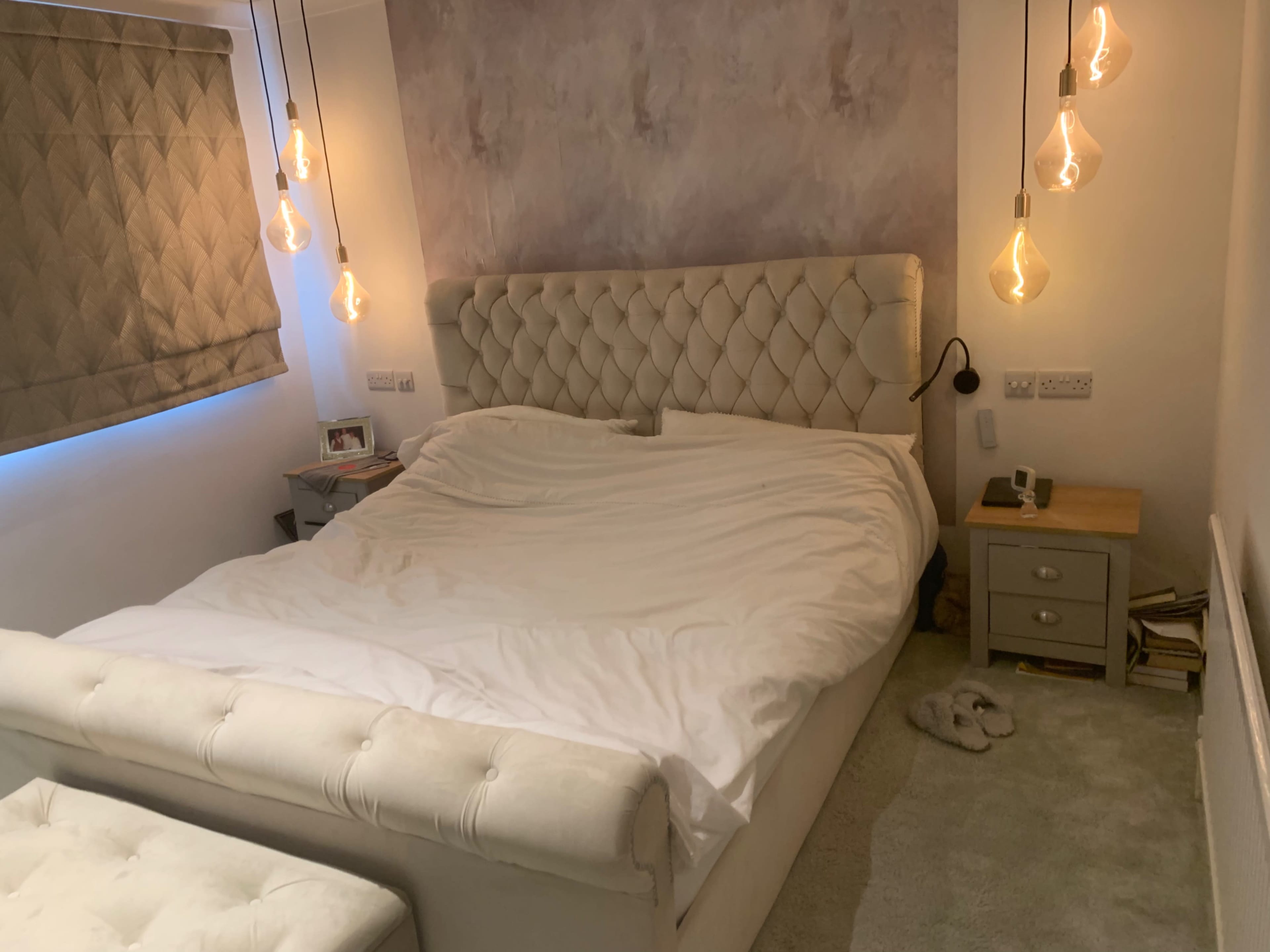 The image shows a bedroom with a large, tufted bed, a bedside table, and pendant lights hanging from the ceiling.