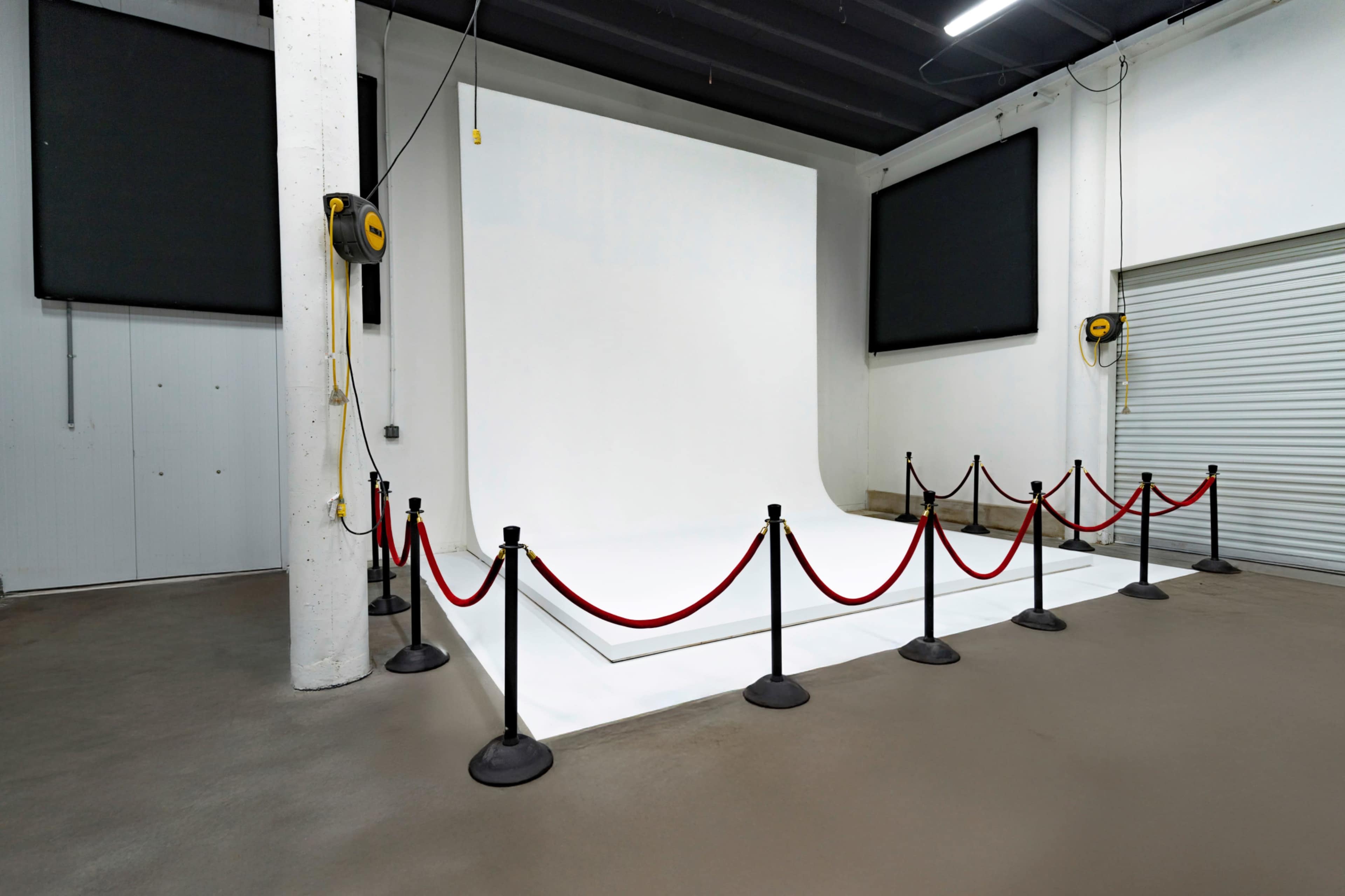 The image shows an empty photography studio featuring a white backdrop, a raised platform, and red ropes with stanchions enclosed around it.