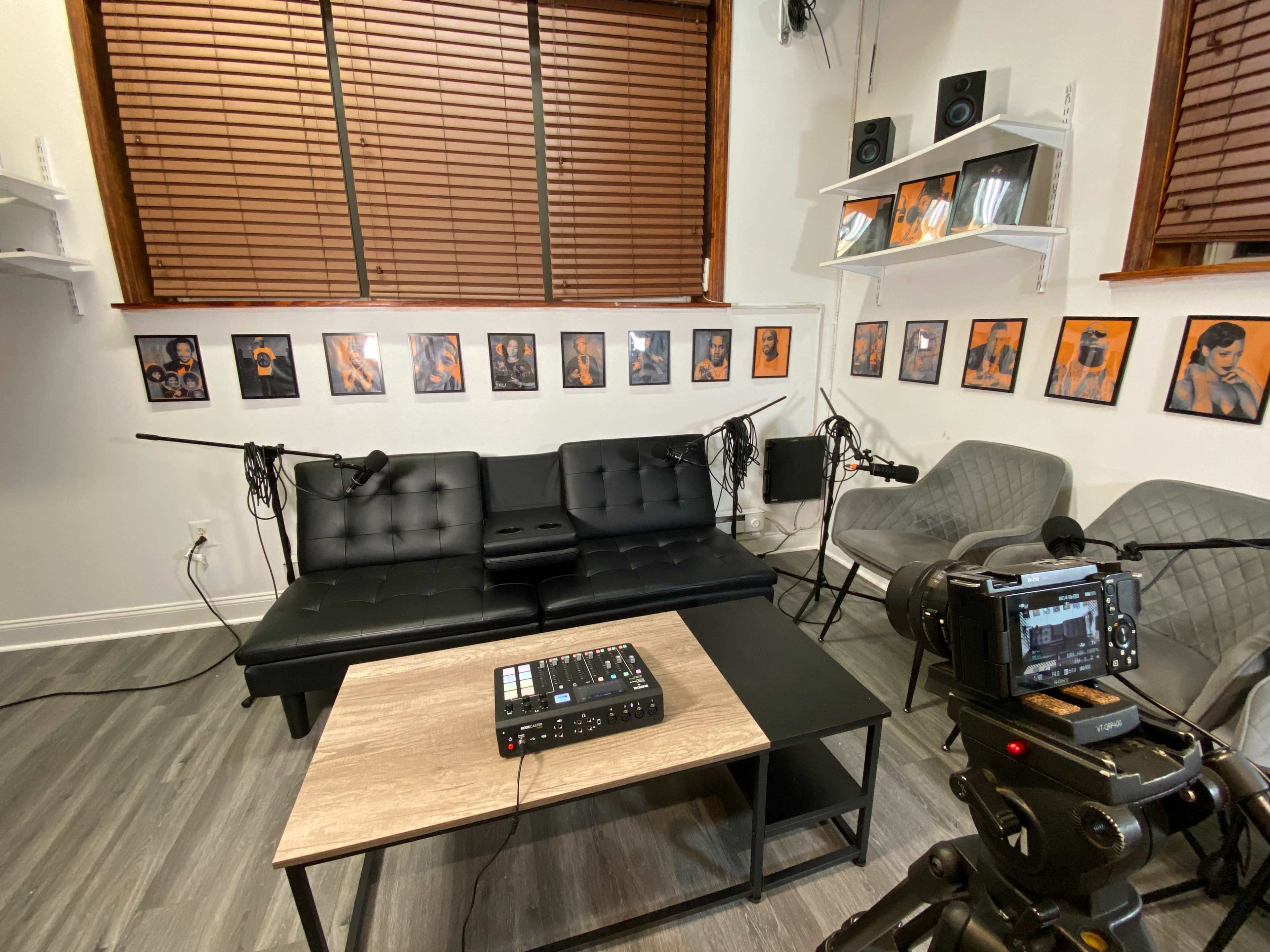 The image shows a recording studio with a black couch, a coffee table, two microphones set up, and framed artwork on the walls.