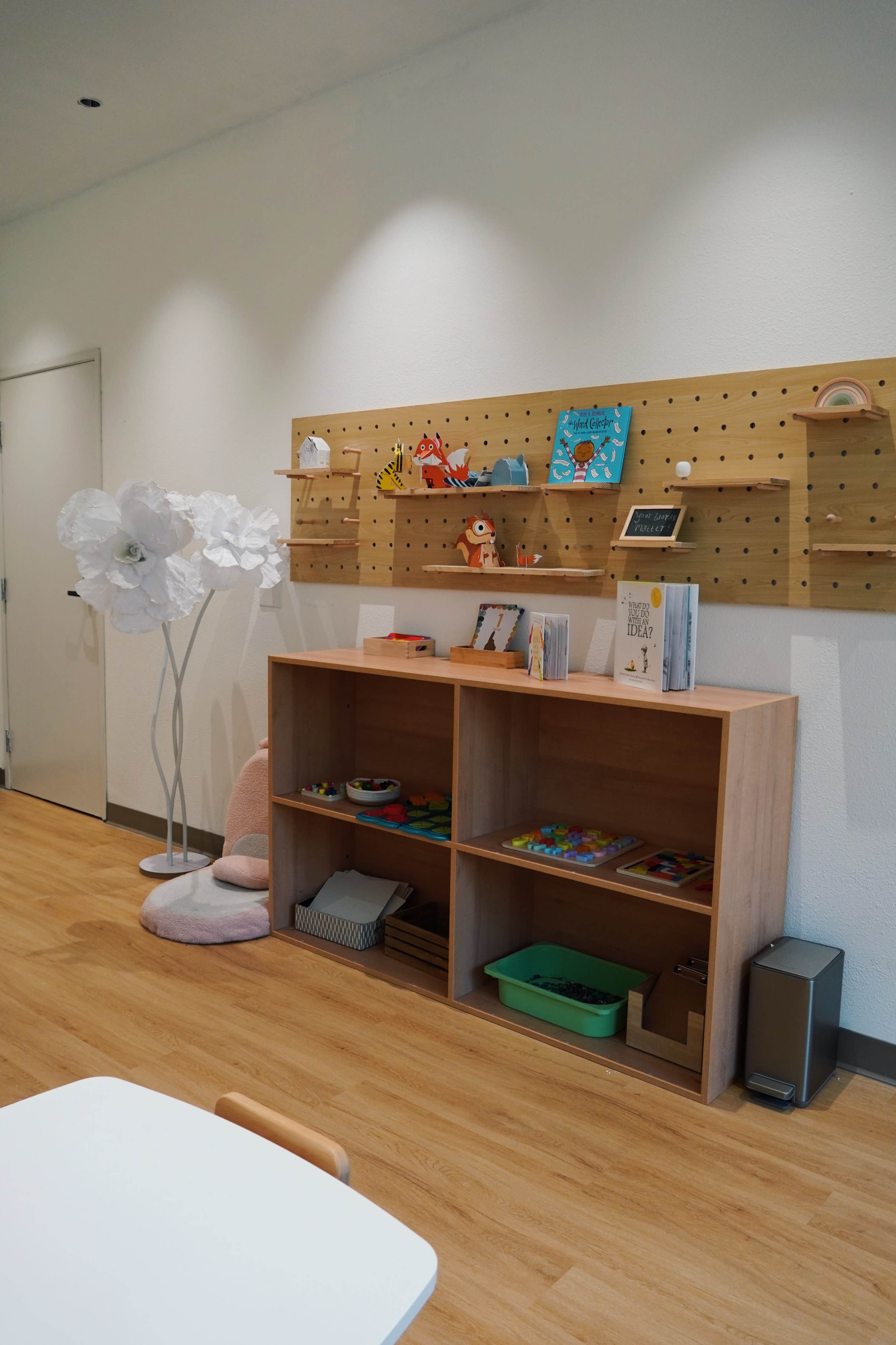 The image shows a children's playroom with a wooden bookshelf, various toys, and a decorative white flower lamp against a white wall.