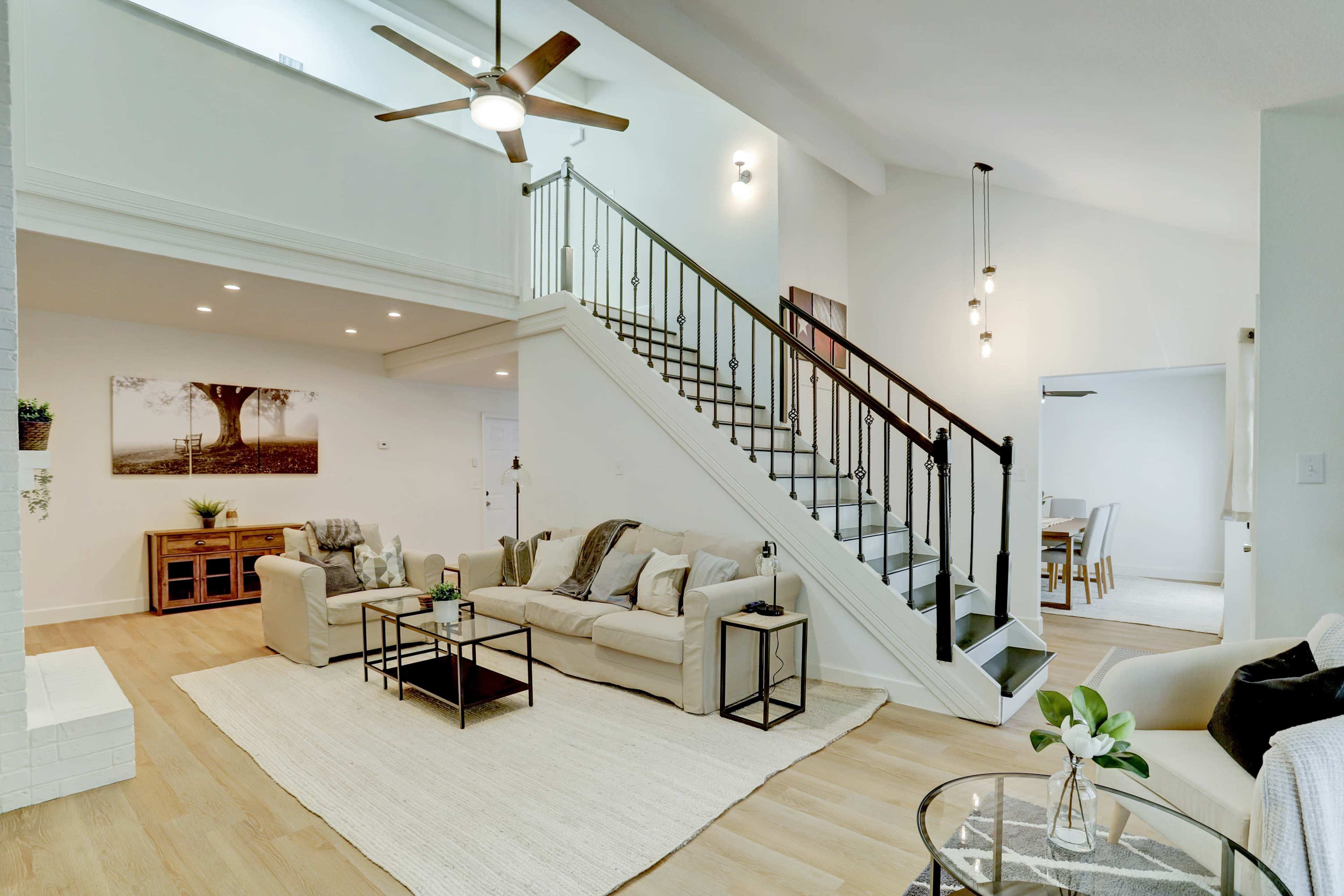 A modern living area with a light-colored sectional sofa, a coffee table, a staircase, and a dining area visible in the background.
