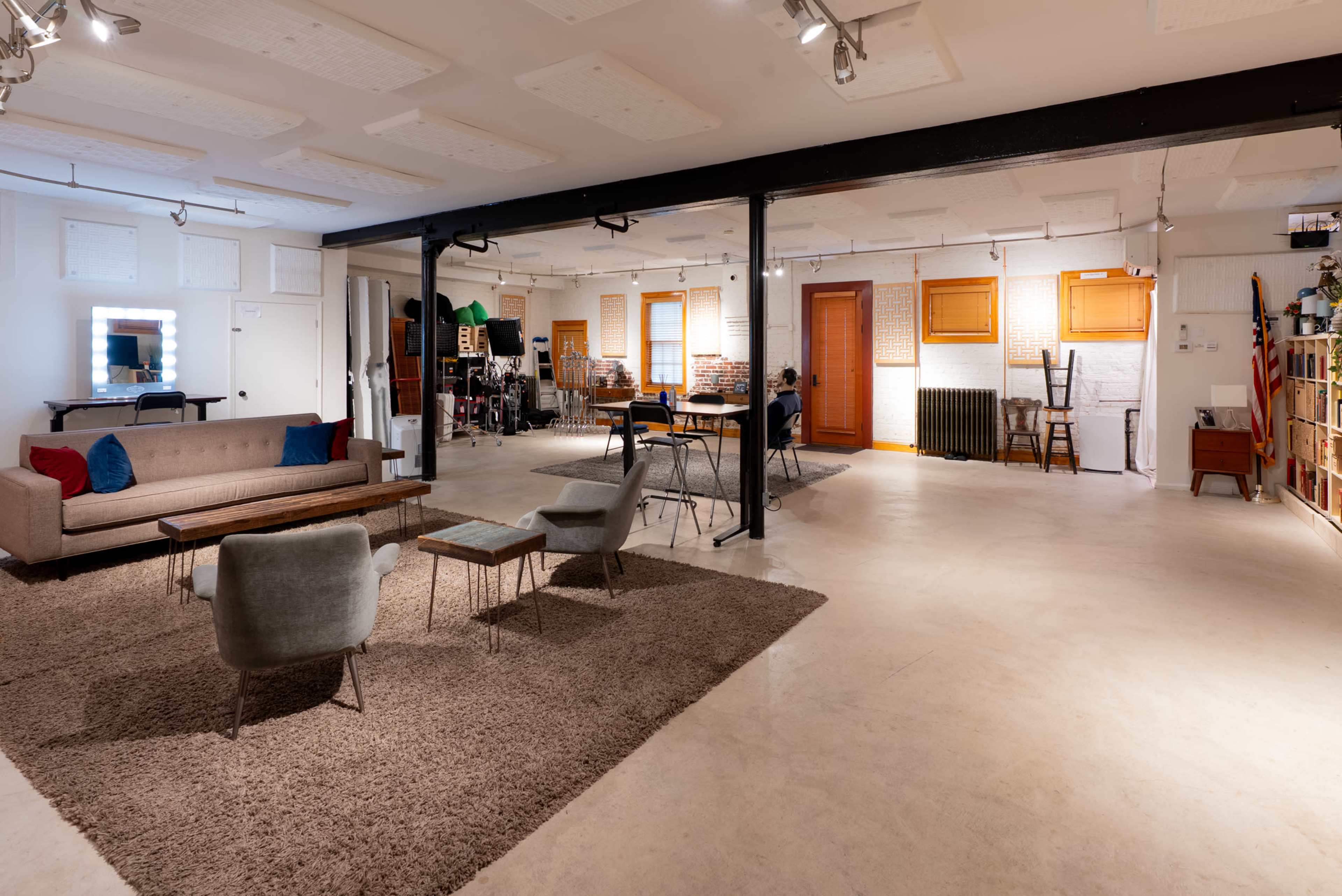 The image shows a spacious, modern studio interior with a seating area, dining table, and various decorative elements against a neutral backdrop.