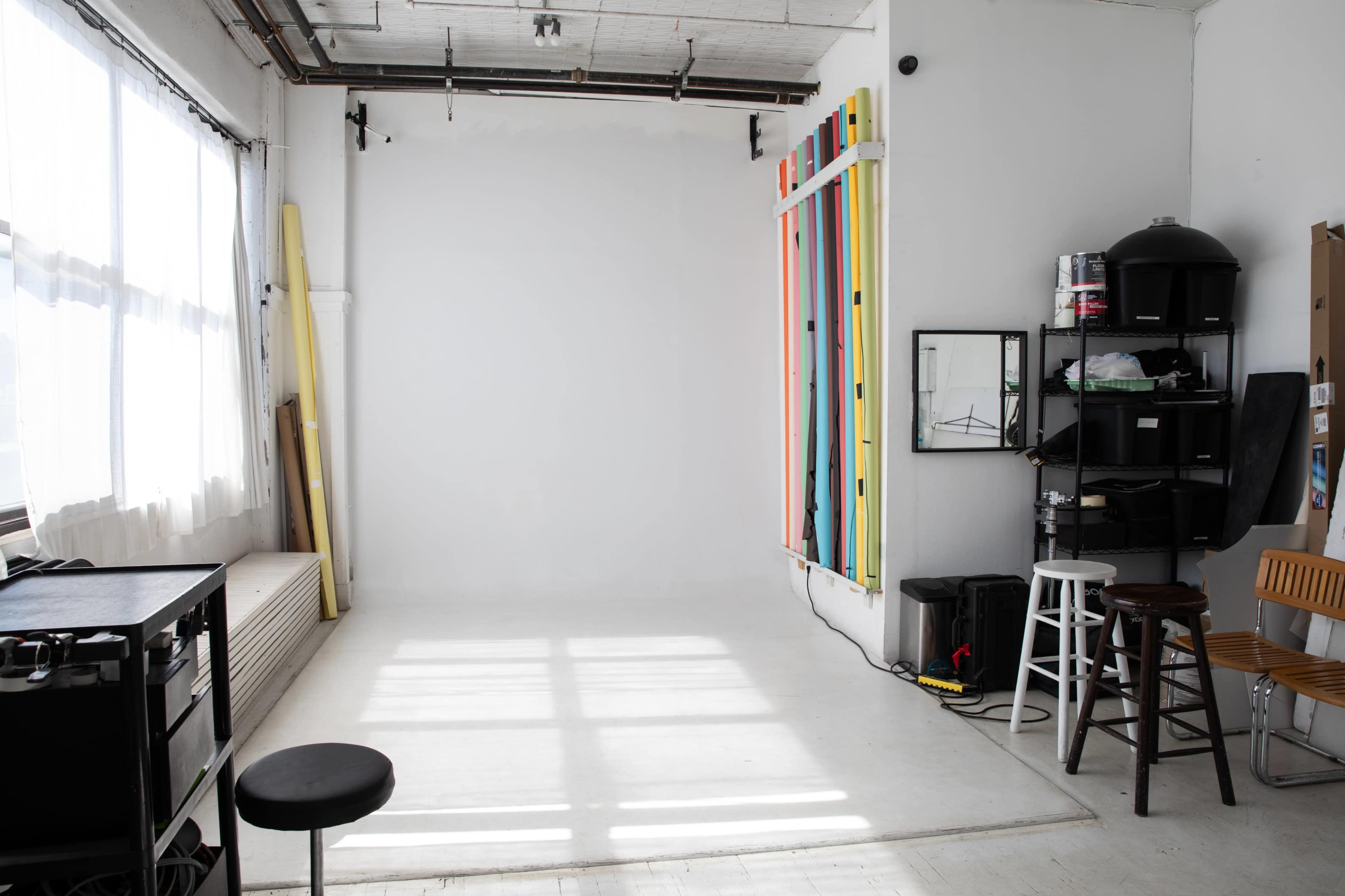The image shows a spacious, well-lit photography studio featuring a white backdrop, colorful vertical panels, and various seating and storage items.