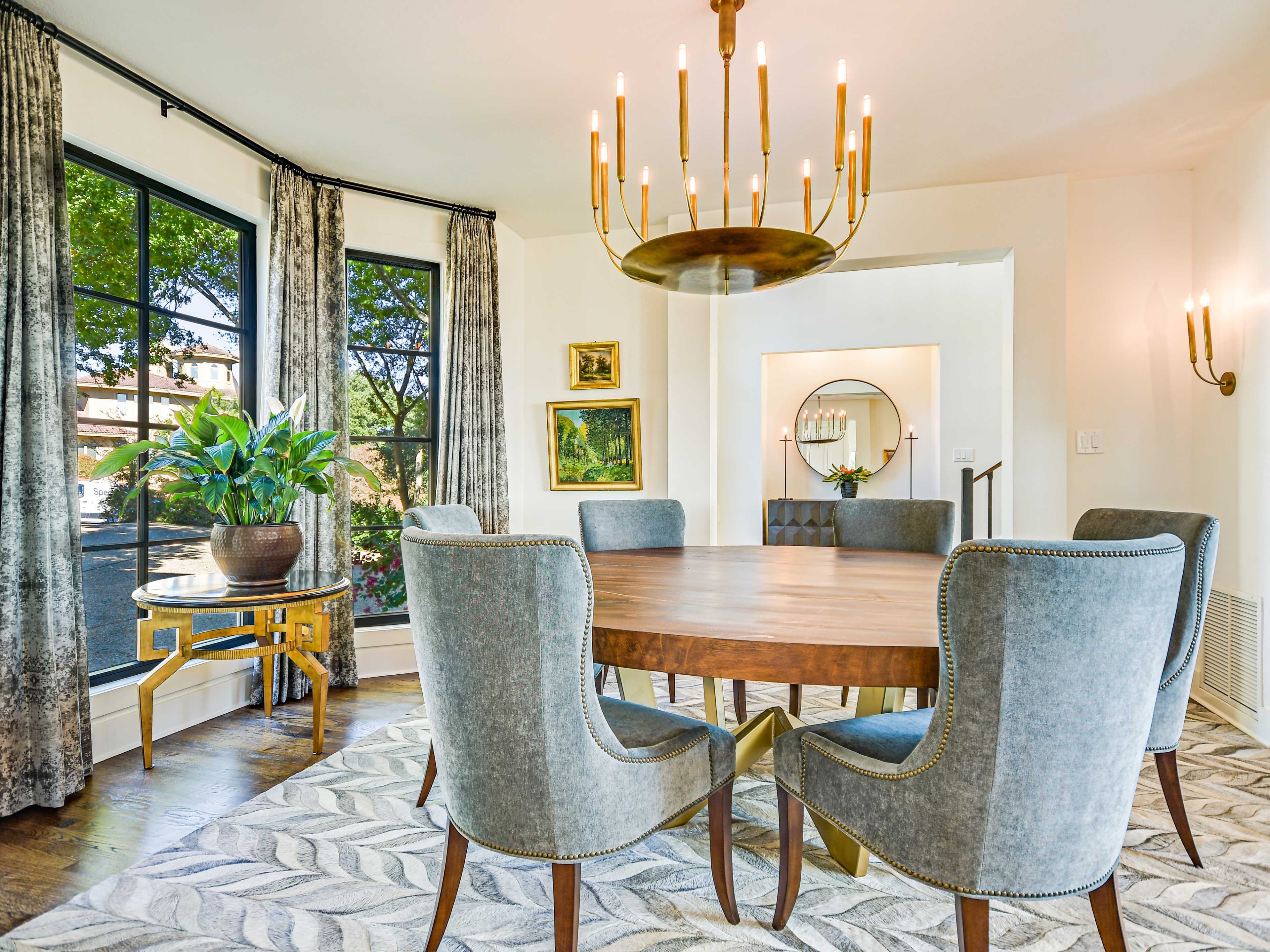 A dining room features a round wooden table surrounded by six upholstered chairs, a chandelier overhead, and large windows with greenery outside.