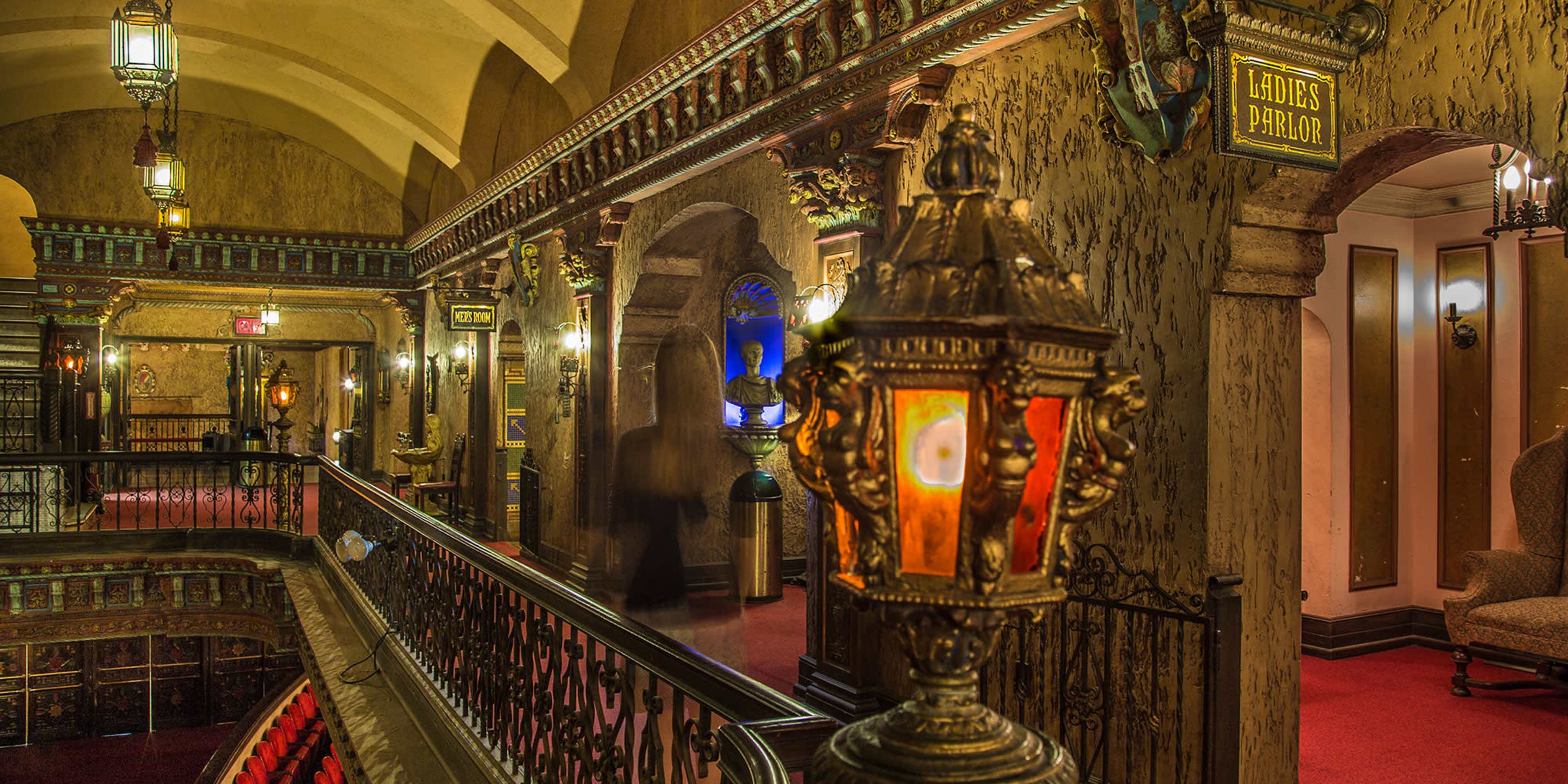 The image shows a vintage theater hallway with ornate details, decorative lighting fixtures, and signs indicating the locations of the ladies' parlor.