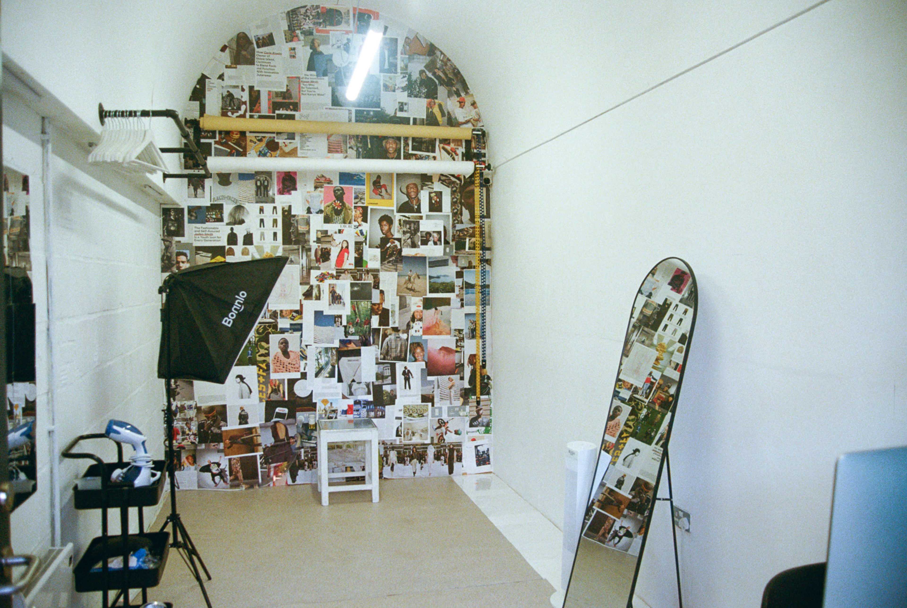 The image shows a minimalist room with white walls adorned with a collage of fashion photographs, a large mirror, and a photography lighting setup.