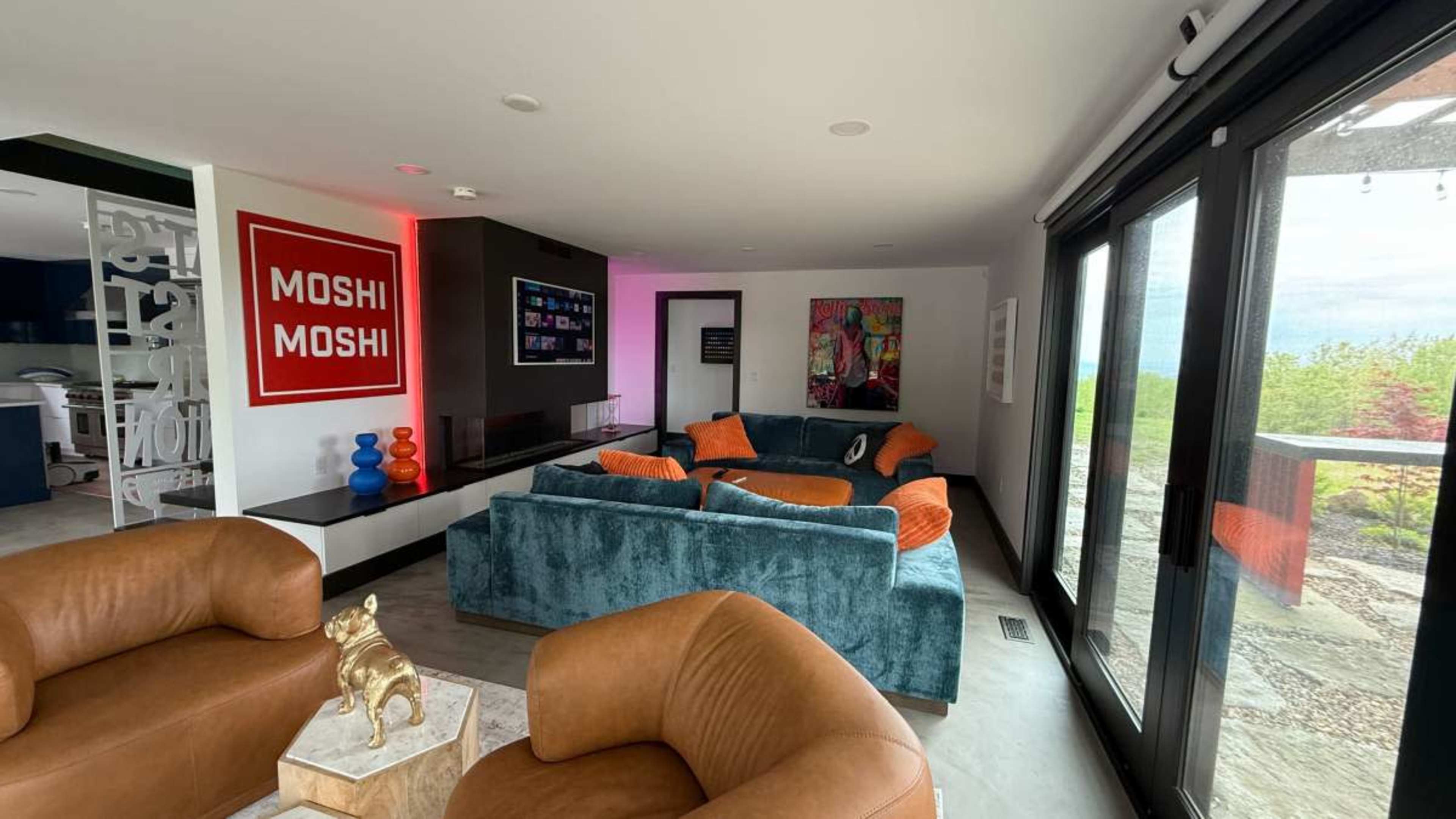 The image shows a modern living room with bright orange and teal furniture, a large window overlooking a garden, and a television on the wall.