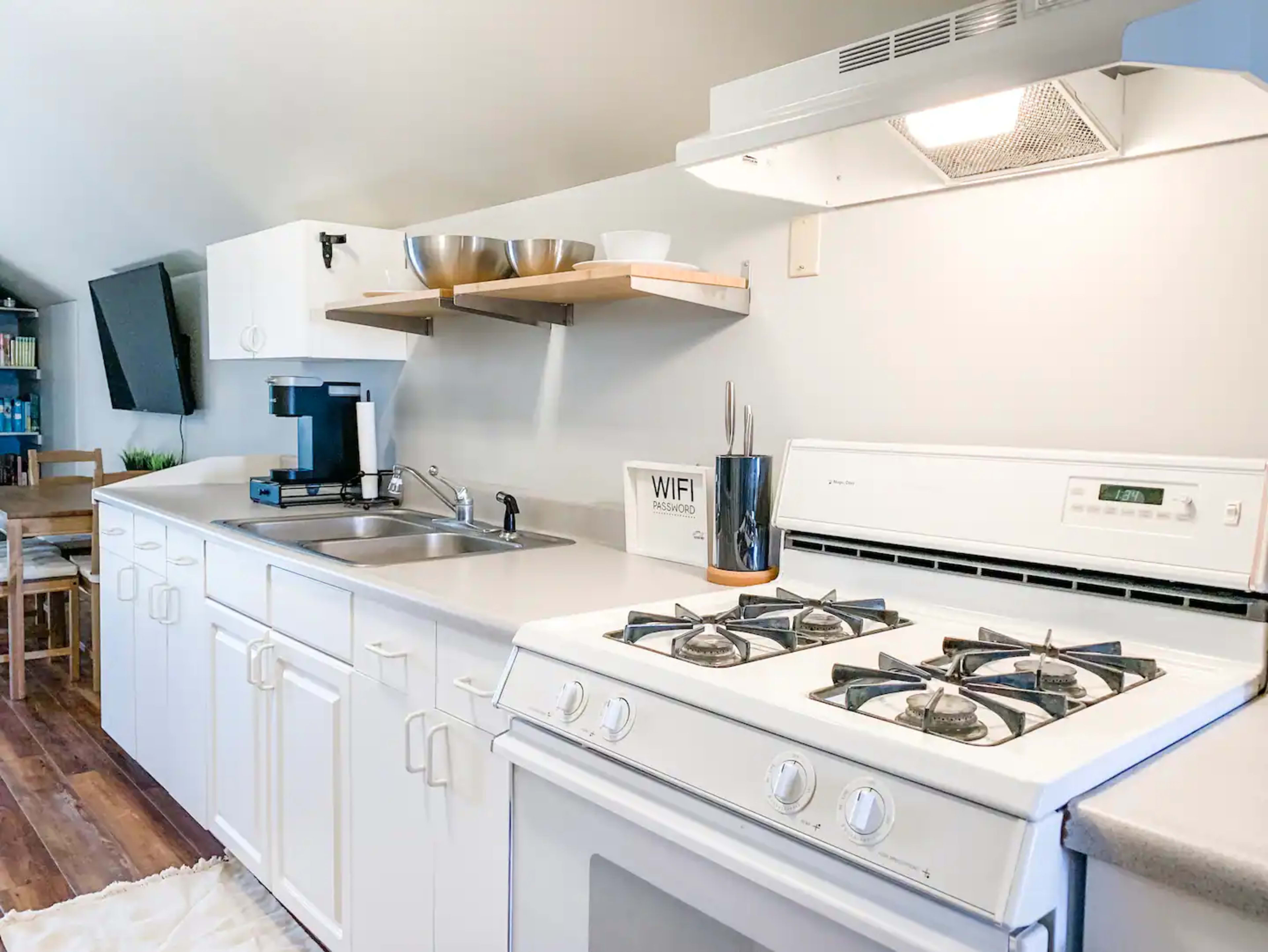 A modern kitchen with white cabinets, a sink, a stove, and a coffee maker on the counter, alongside open shelving with bowls and a decorative sign.