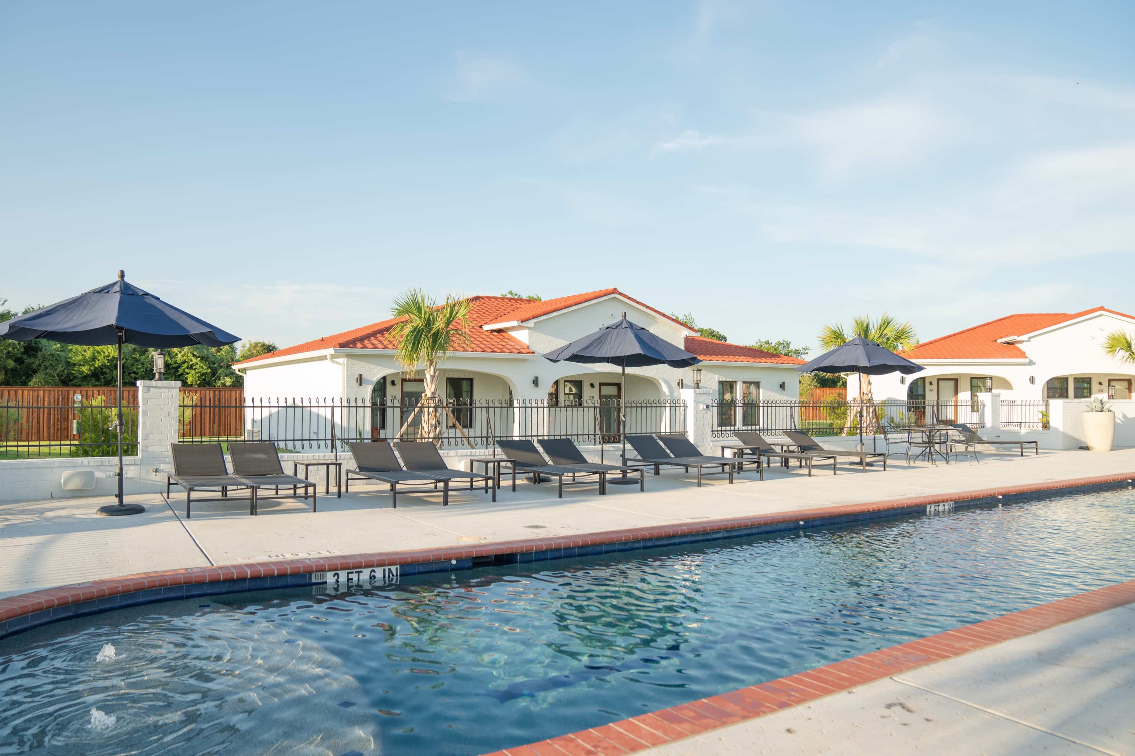 A swimming pool with lounge chairs and umbrellas is situated beside a white building with a red-tiled roof.