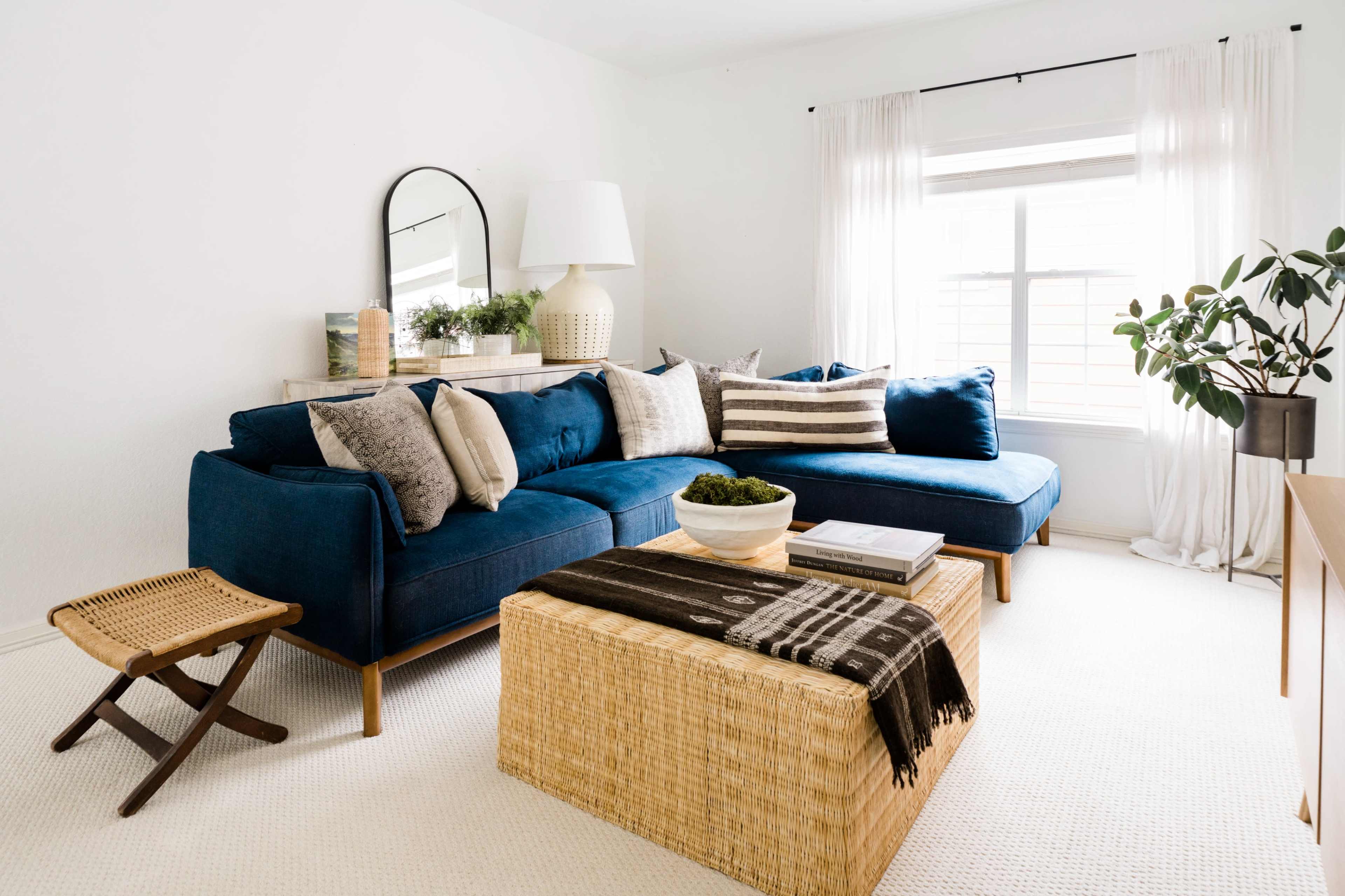 The living room features a blue sectional couch, a woven coffee table, and a potted plant beside a window with sheer curtains.