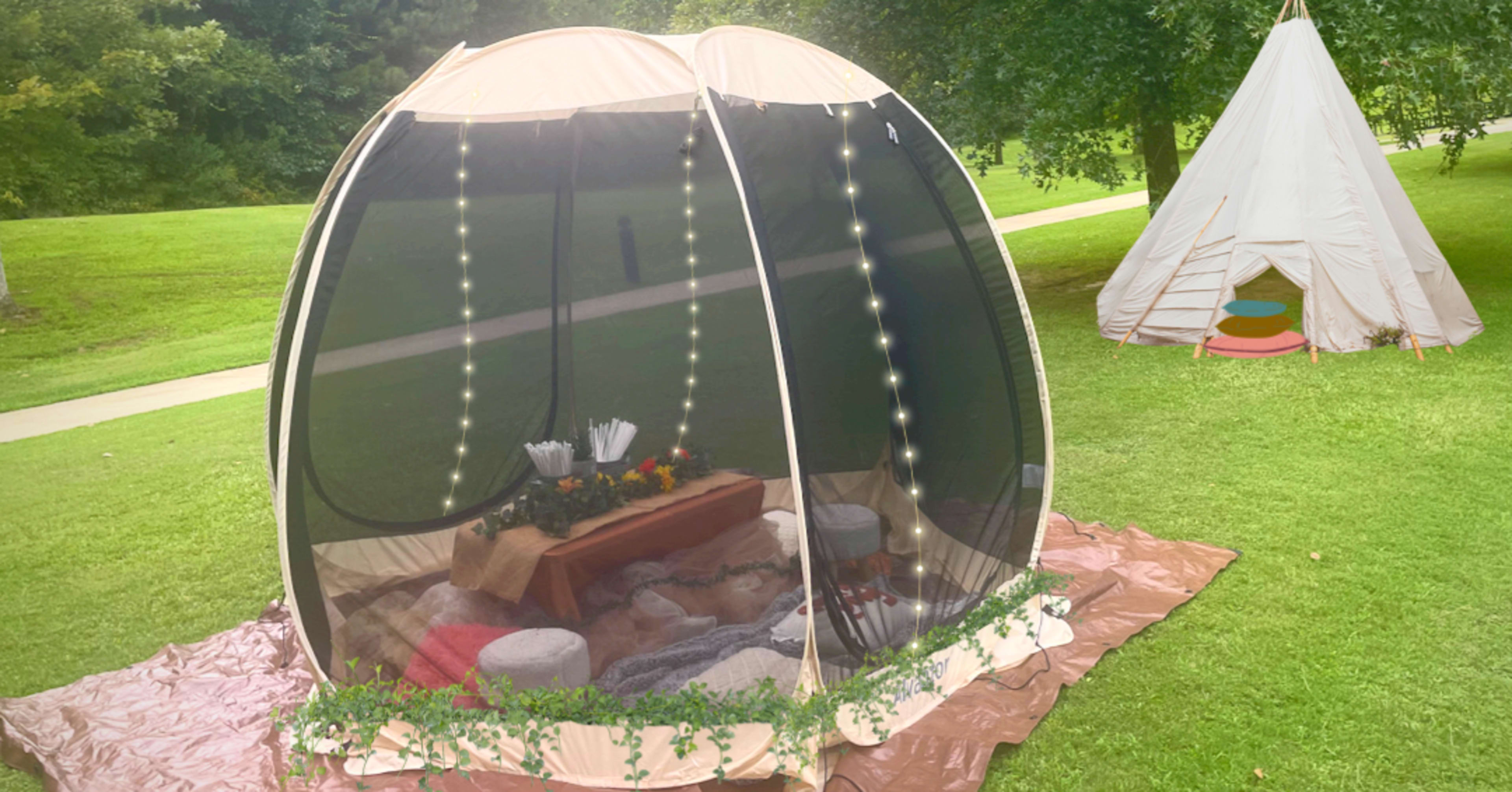 A large, mesh tent filled with decorations and seating is set up on a grassy area alongside a smaller, traditional teepee.