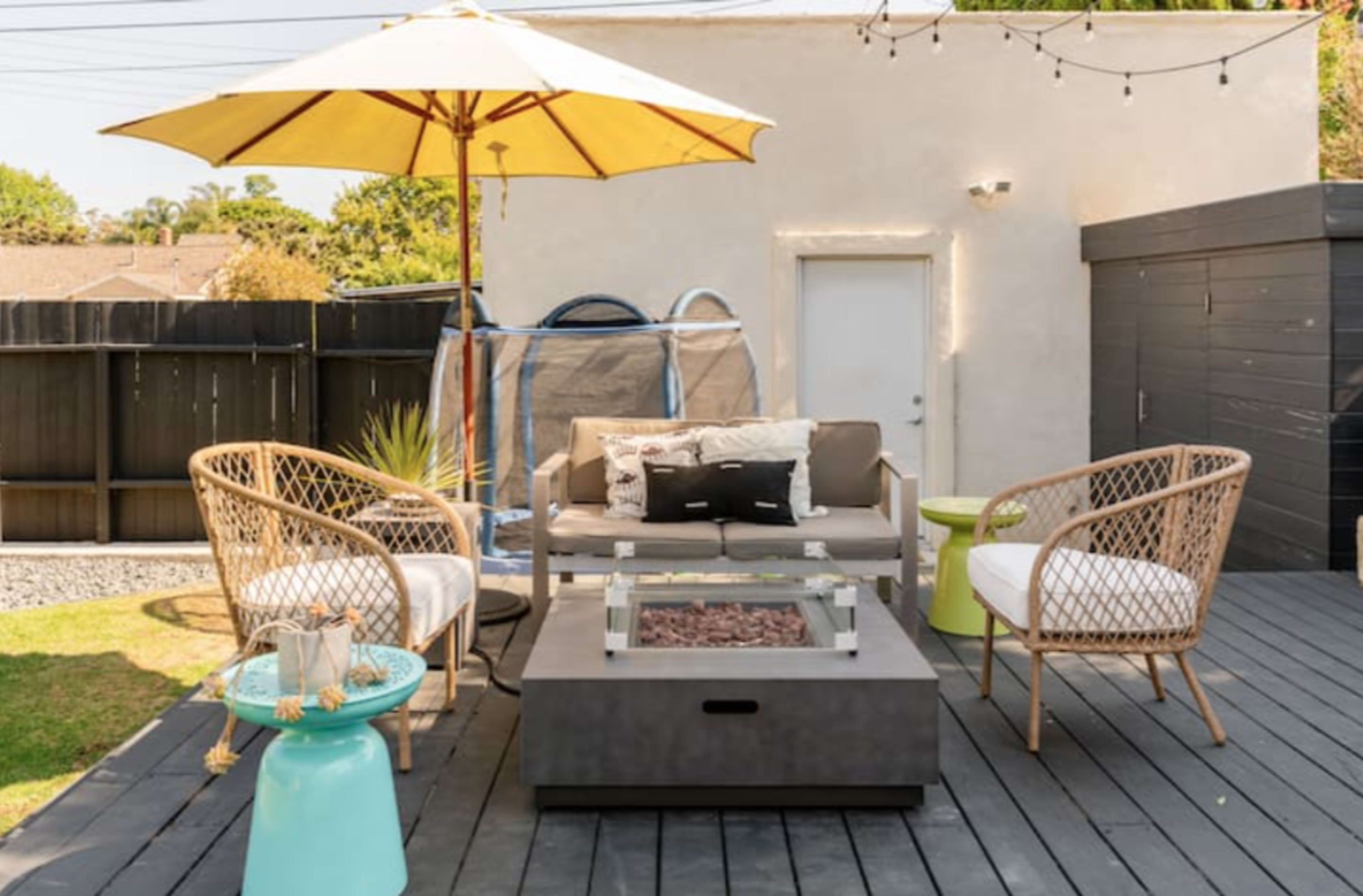 A patio area featuring a couch with an umbrella, two woven chairs, a coffee table with a fire pit, and a trampoline in the background.