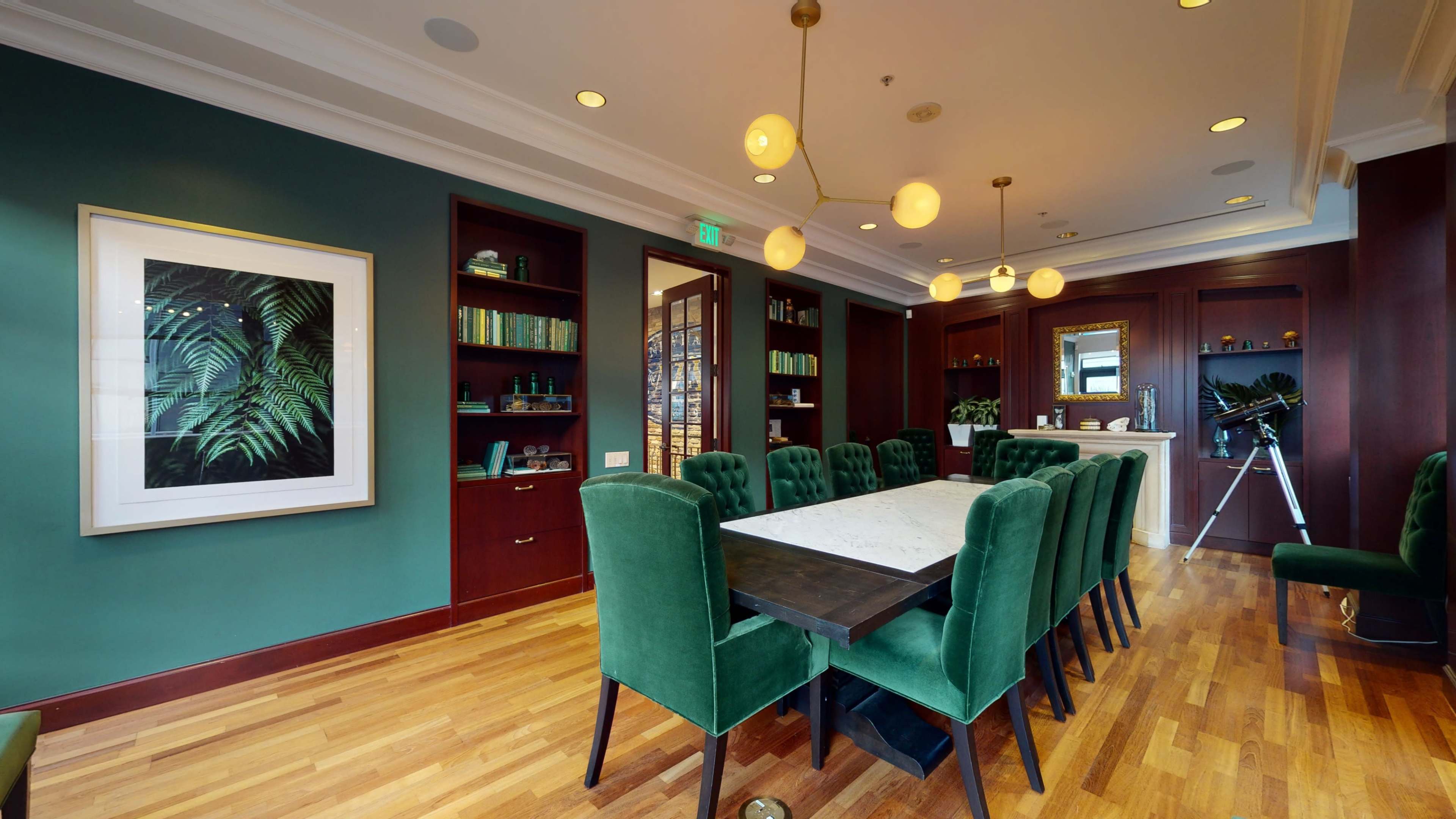 A conference room with a long black table surrounded by green upholstered chairs, framed by dark wood shelves and a large window.