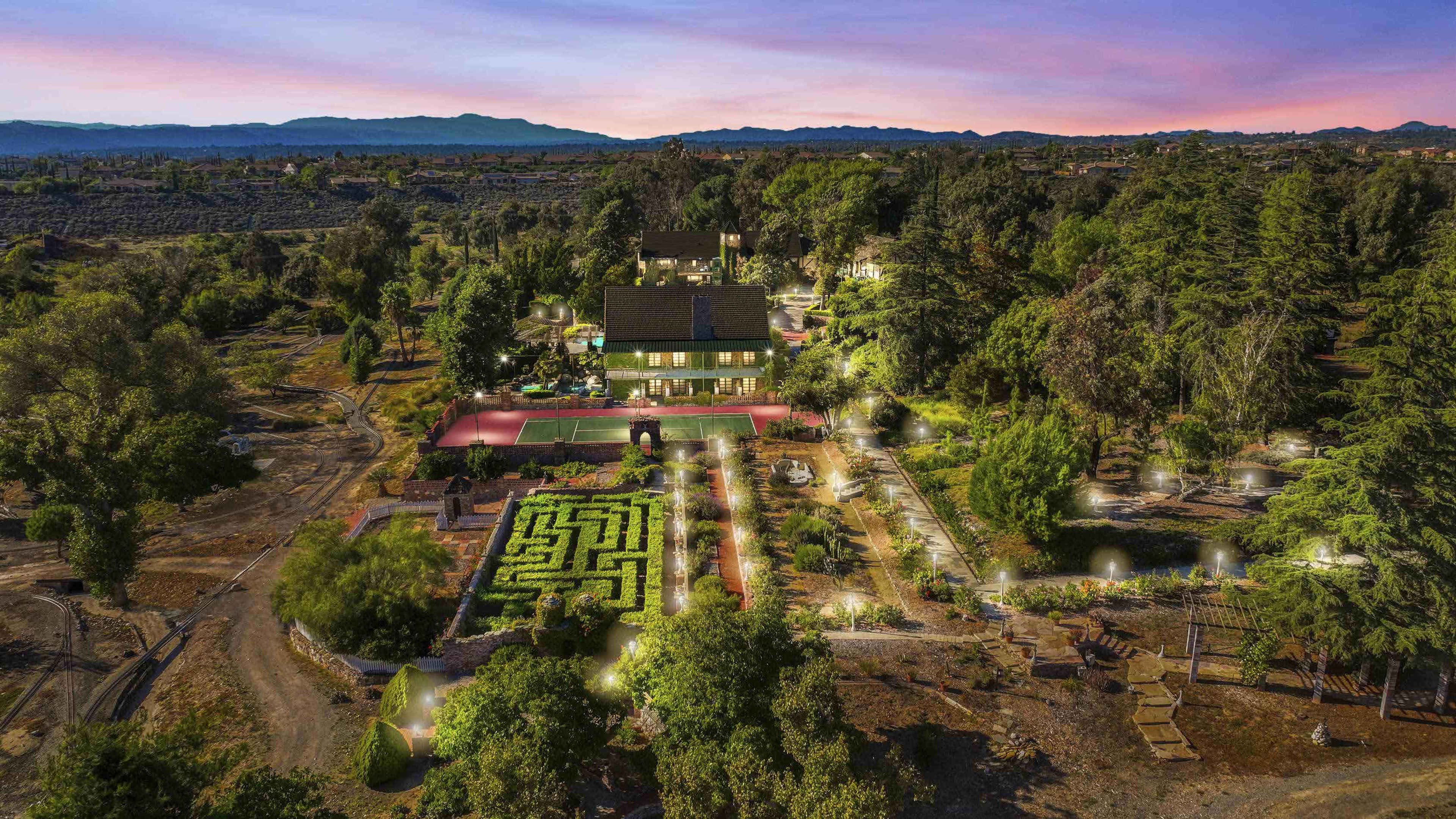 The image shows a sprawling estate featuring a maze garden, tennis court, and surrounding greenery under a sunset sky.