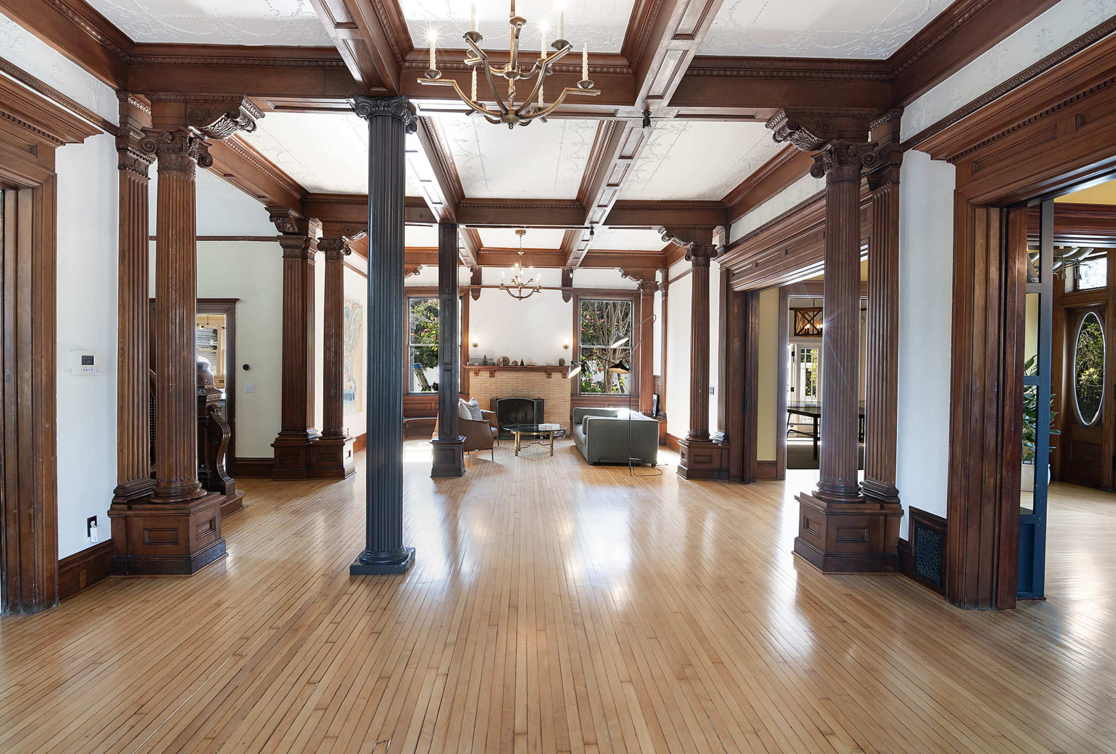 The image shows a spacious room with polished wooden floors, tall columns, and a central seating area featuring a sofa and a fireplace.