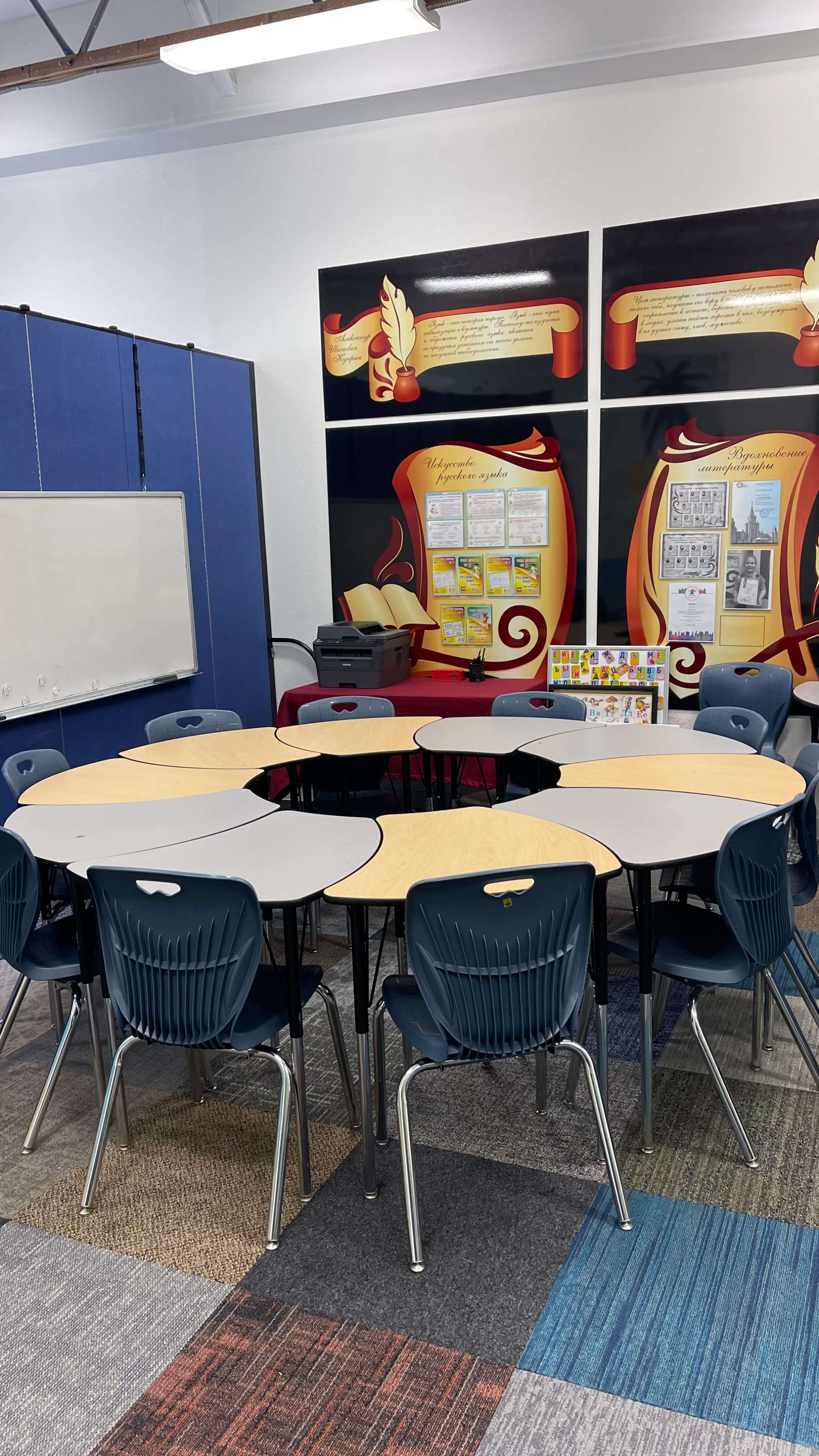 A circular arrangement of tables with blue chairs in a classroom featuring colored wall displays and a whiteboard.