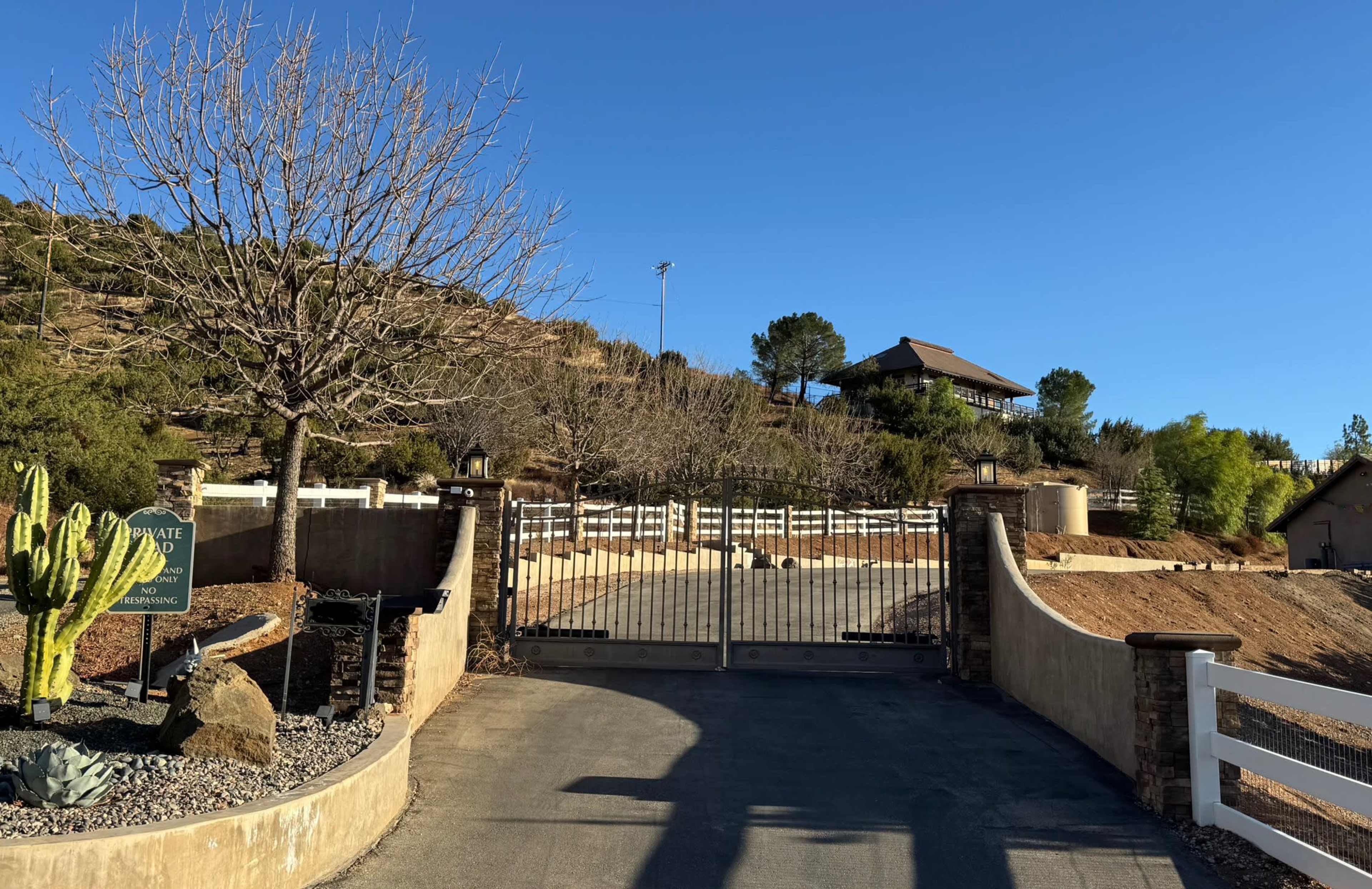 A gated entrance leads to a property with a house on a hill, surrounded by sparse vegetation and blue sky.