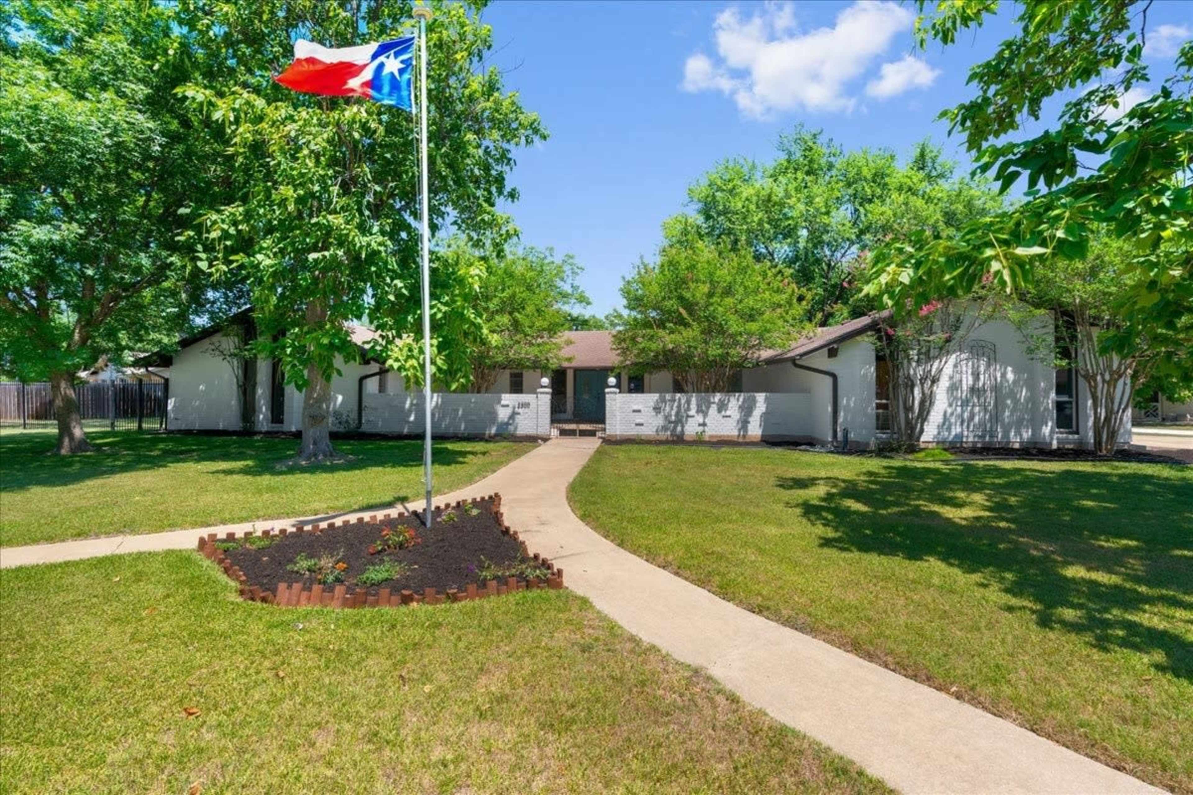 A single-story house with a Texas flag on a flagpole in the front yard, surrounded by green grass and trees.