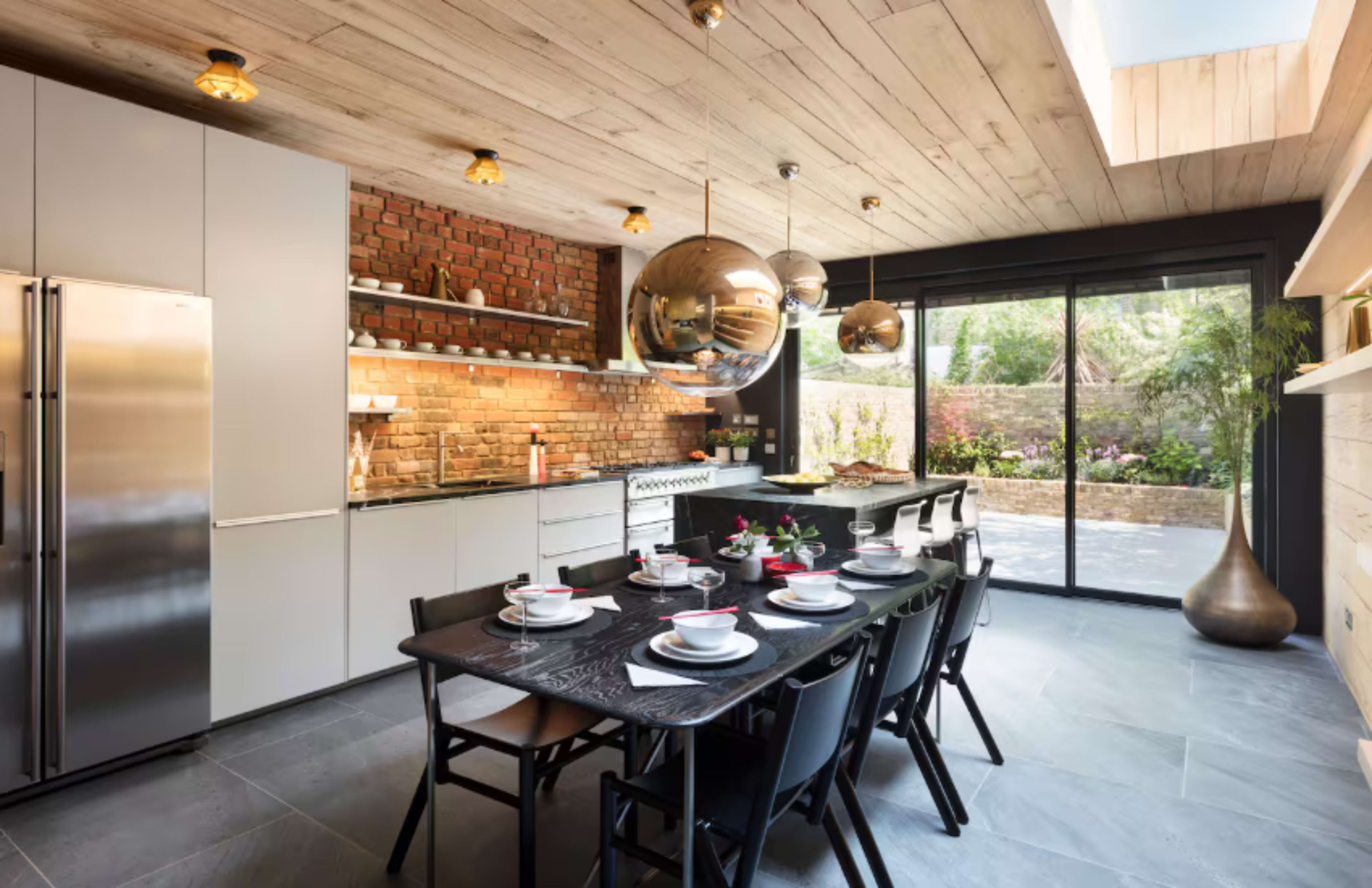 A modern kitchen features sleek cabinetry, a brick wall, and a dining table set for a meal, with large glass doors opening to an outdoor garden.