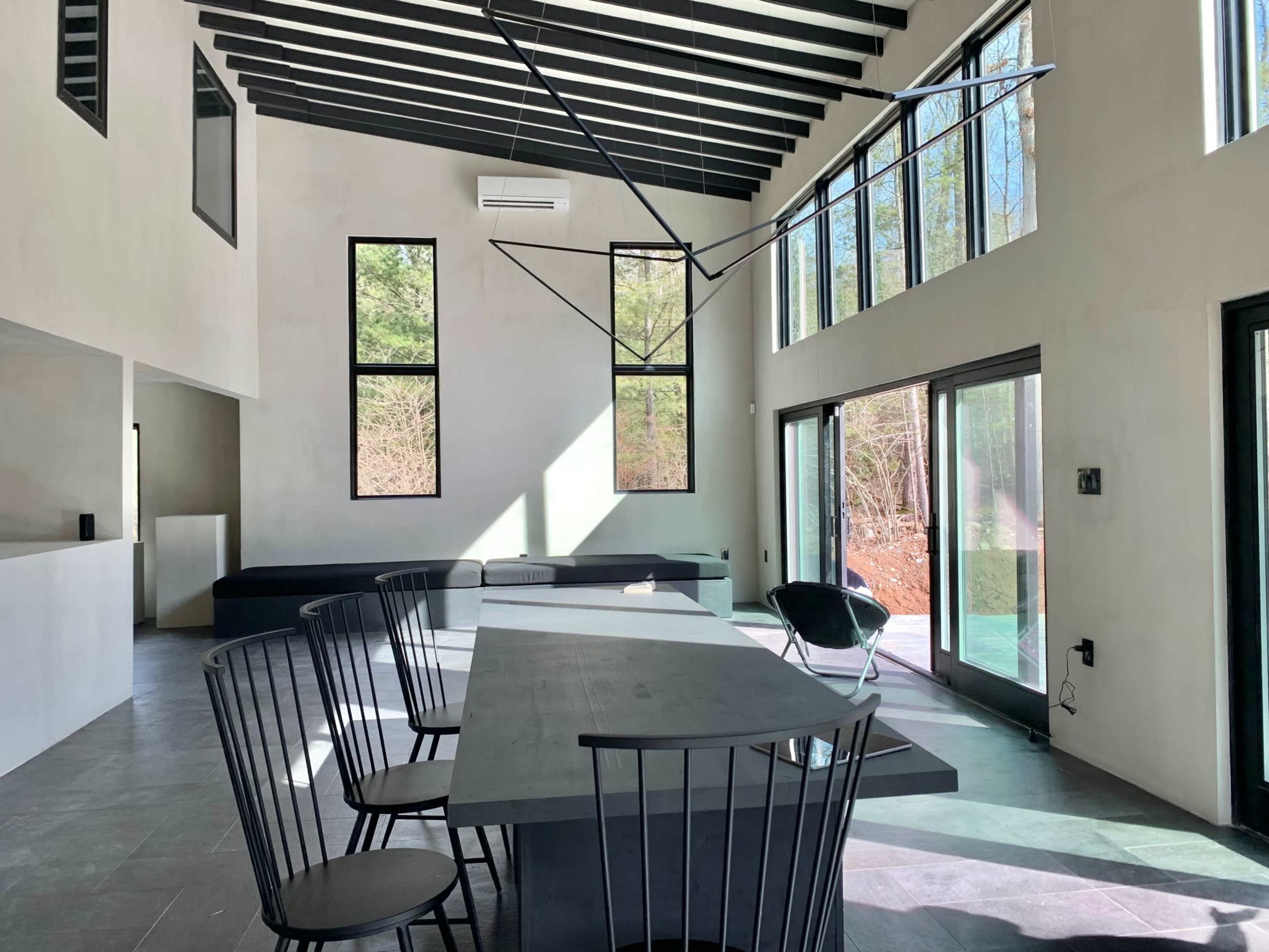 The interior of a modern, open-plan dining area features a large table surrounded by black chairs, with tall windows that allow sunlight to enter and a view of the outdoors.