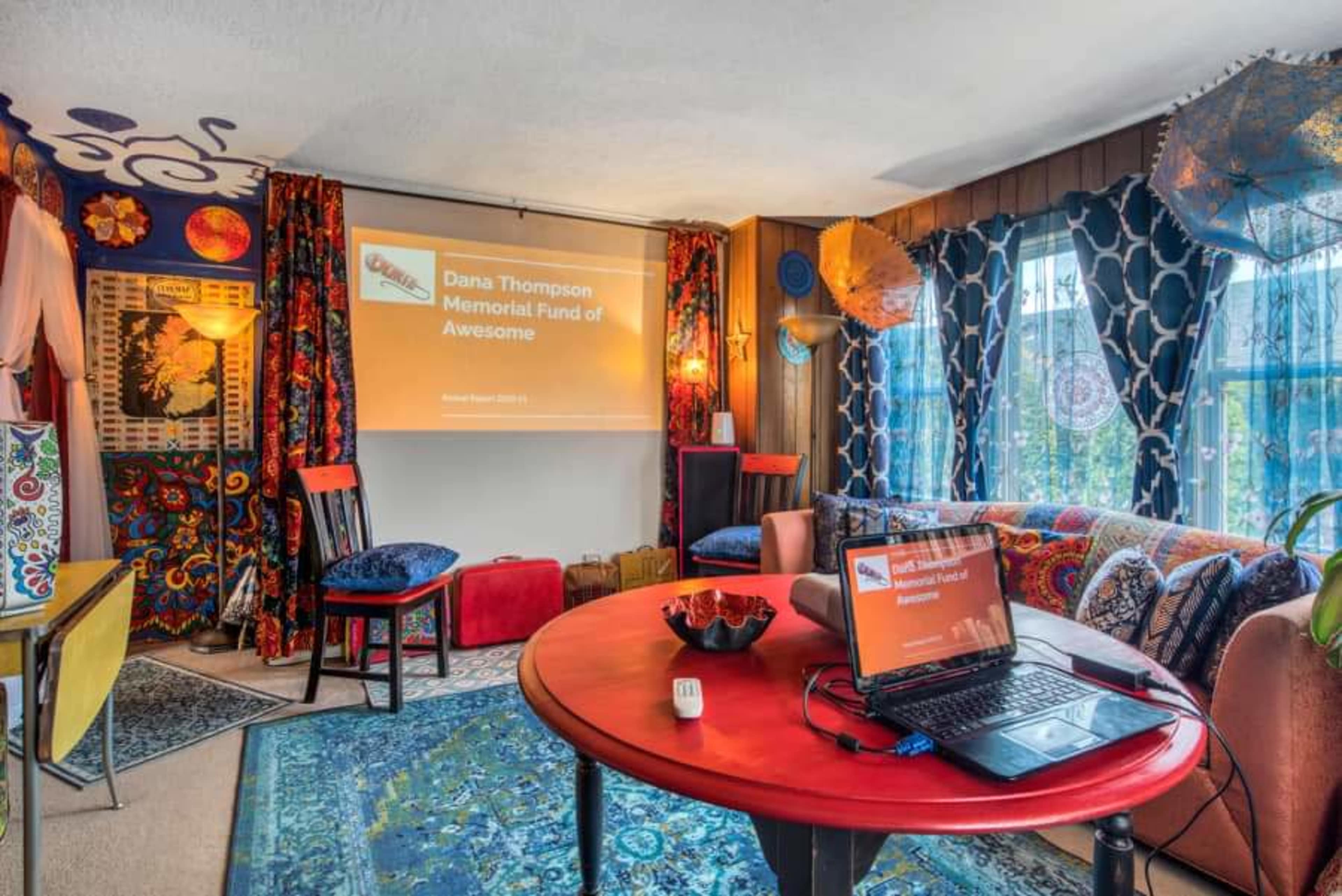A cozy room with colorful decor, featuring a projector screen displaying a presentation, and a laptop on a table surrounded by chairs and a patterned rug.