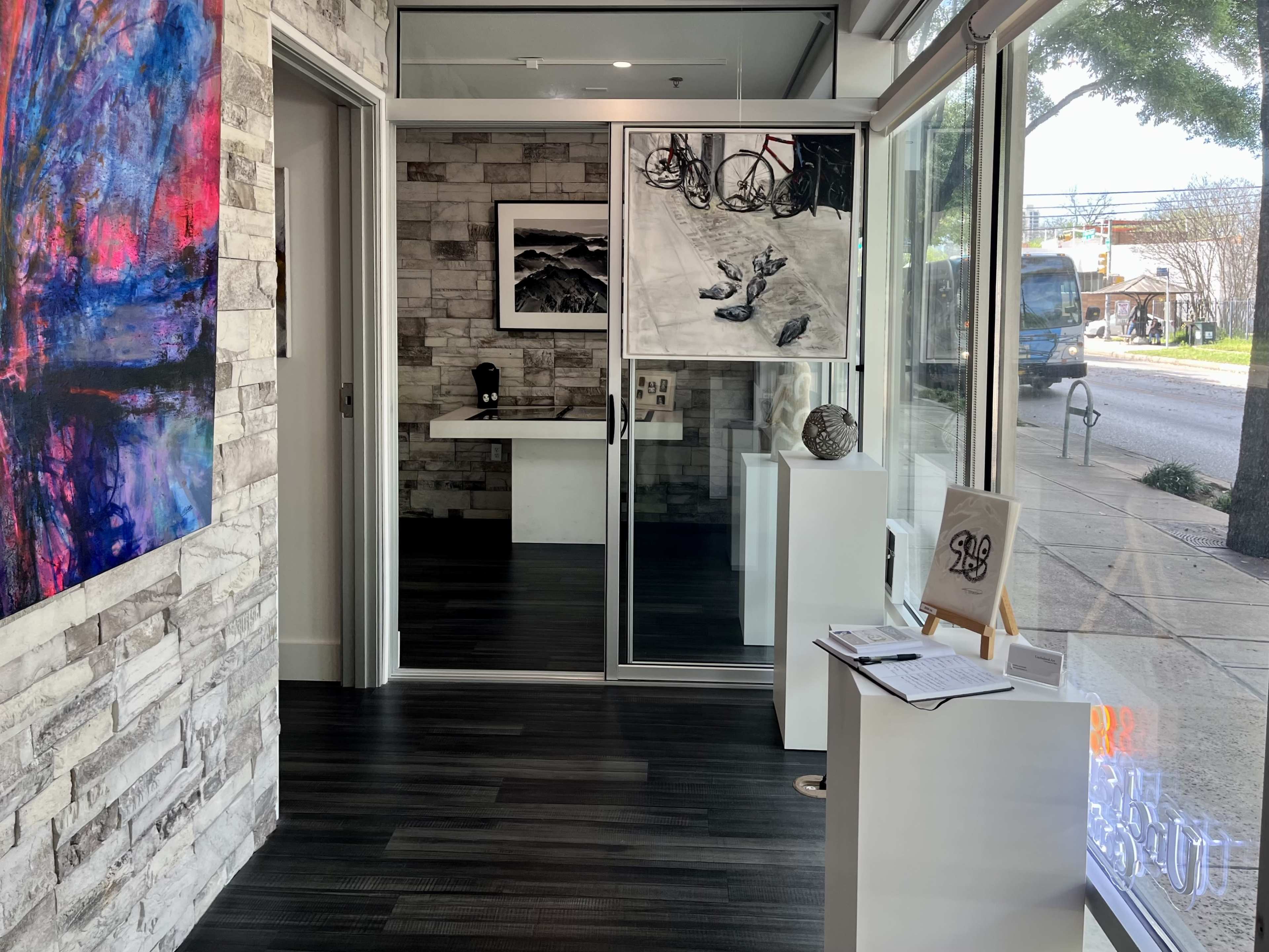 A modern gallery entrance featuring stone walls, a large artwork on the left, and a display area with white pedestals and a sign-in sheet near the doorway.