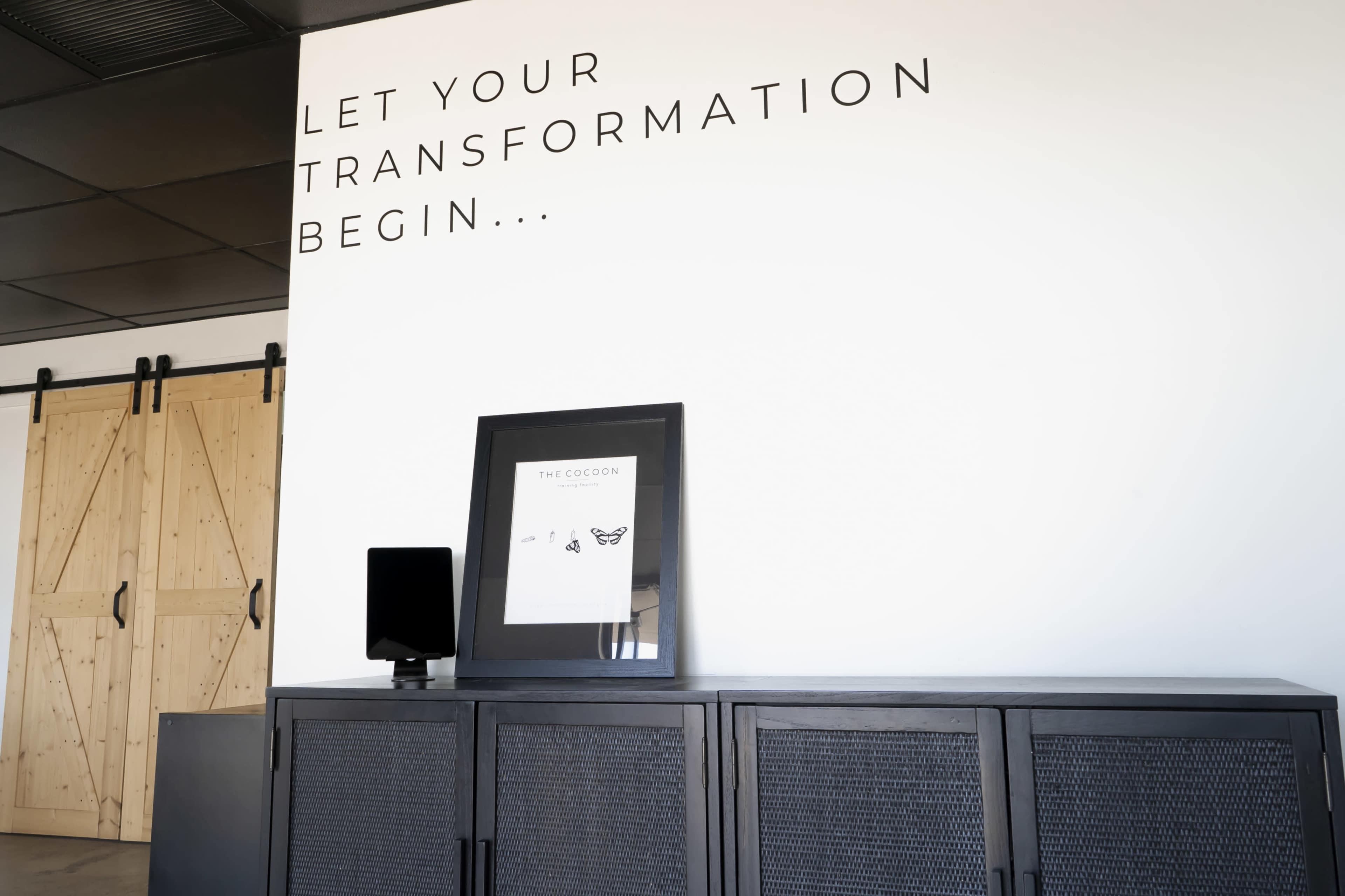 The image shows a modern interior space with a motivational quote on the wall, a framed artwork on a black cabinet, and sliding wooden doors in the background.